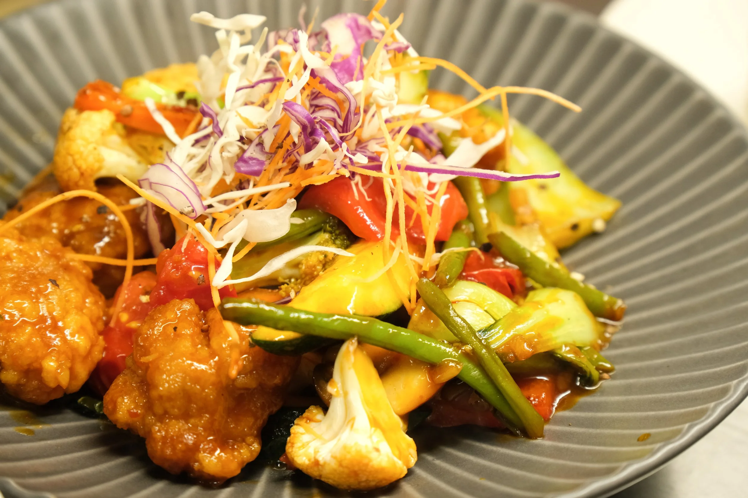A plate of stir-fried vegetables including cauliflower, carrots, green beans, zucchini, and red bell peppers, topped with shredded cabbage and carrots.