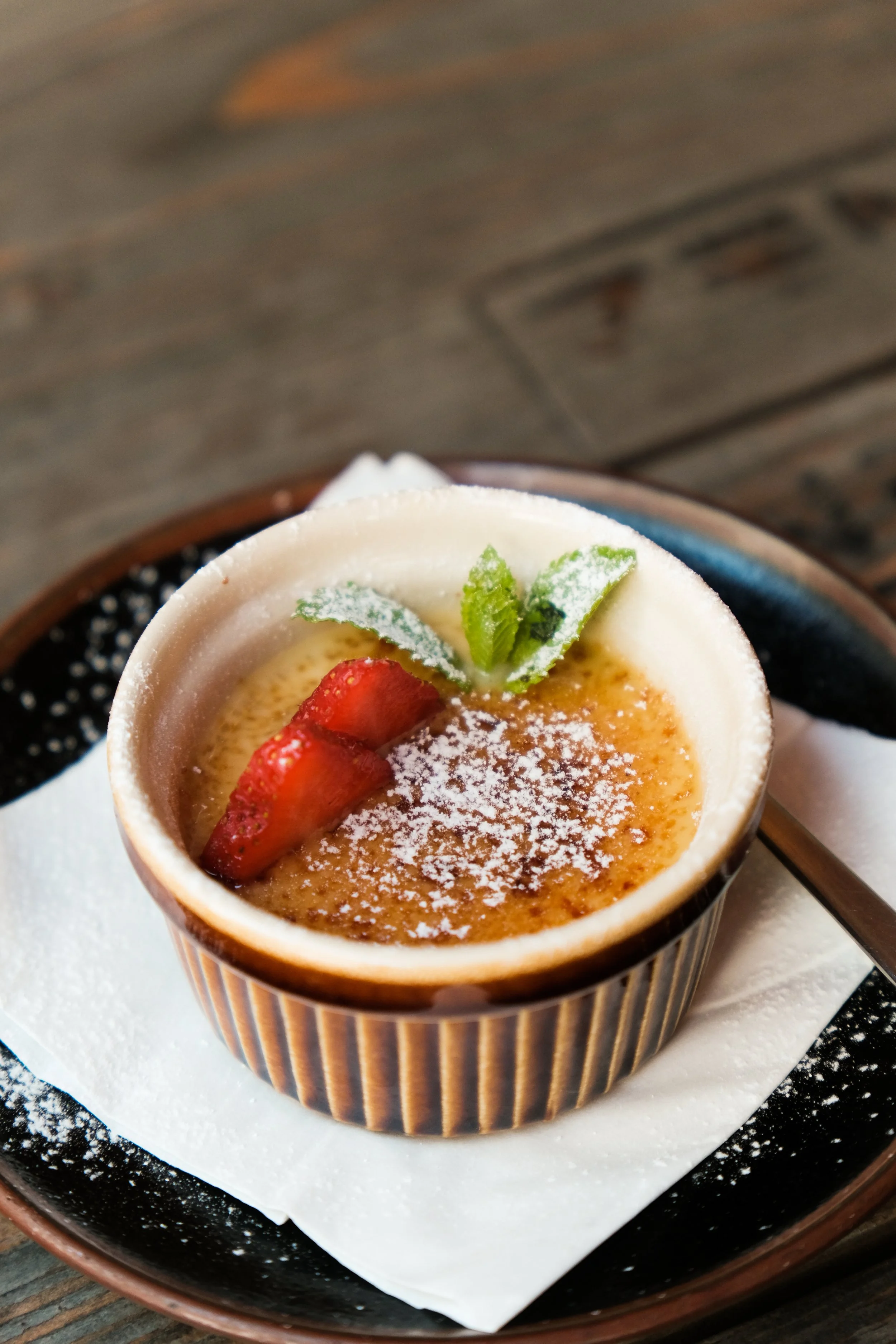Creme brulee topped with strawberries, a mint leaf, and powdered sugar on top of a white napkin, served in a small ramekin.
