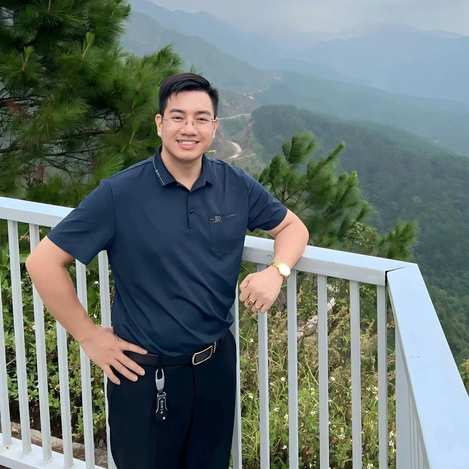 a smiling man with glasses in a navy blue shirt and black pants standing by a white railing on a mountain overlook, with lush green trees and distant mountains in the background.
