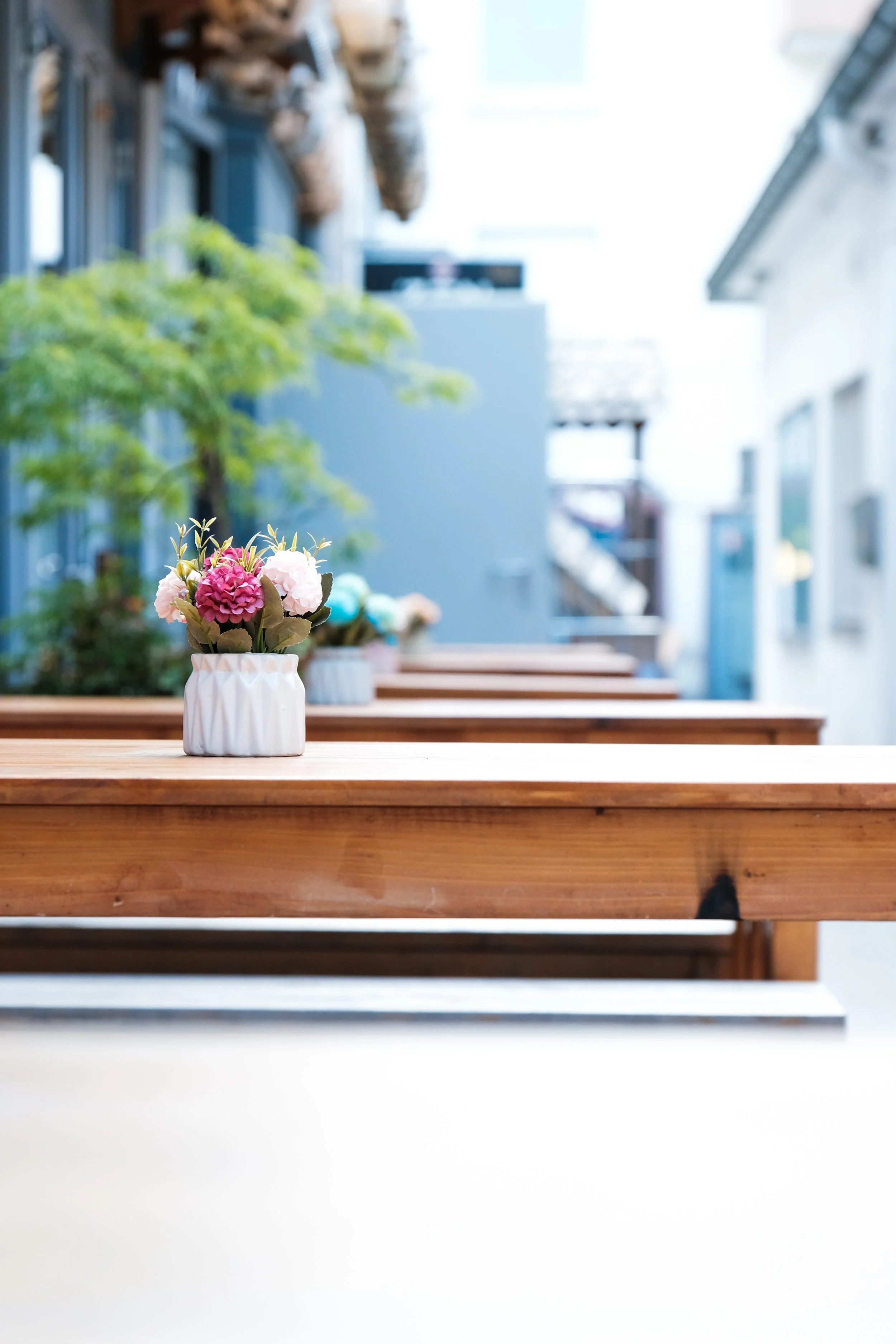 Outdoor cafe table with small pink and white flower arrangement in a white ceramic vase, with a blurred background of similar tables and buildings.