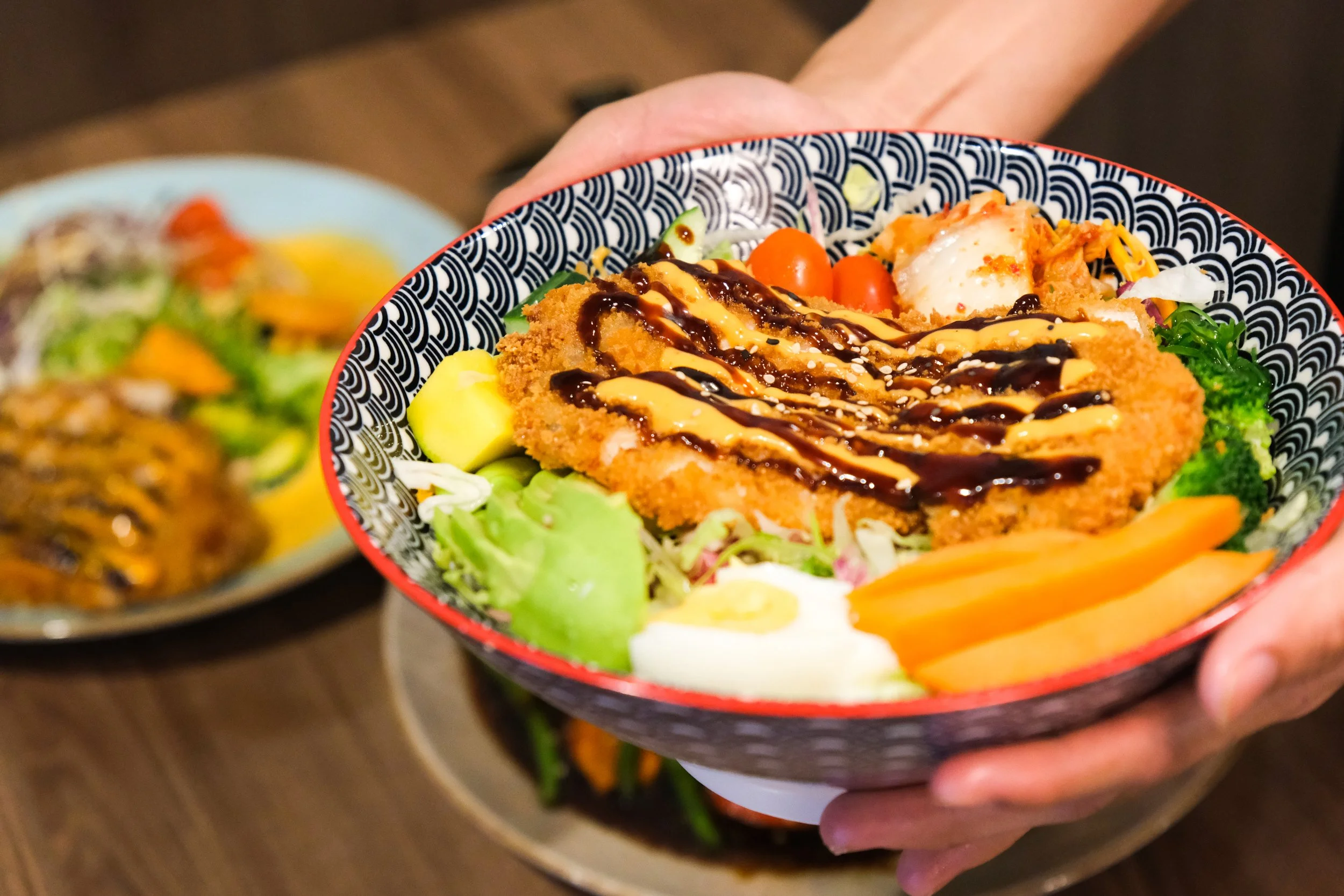 A hand holding a bowl of salad topped with breaded fried meat, drizzled with dark and creamy sauces, with varied vegetables including lettuce, cherry tomatoes, carrot slices, cucumbers, broccoli, and boiled eggs visible.