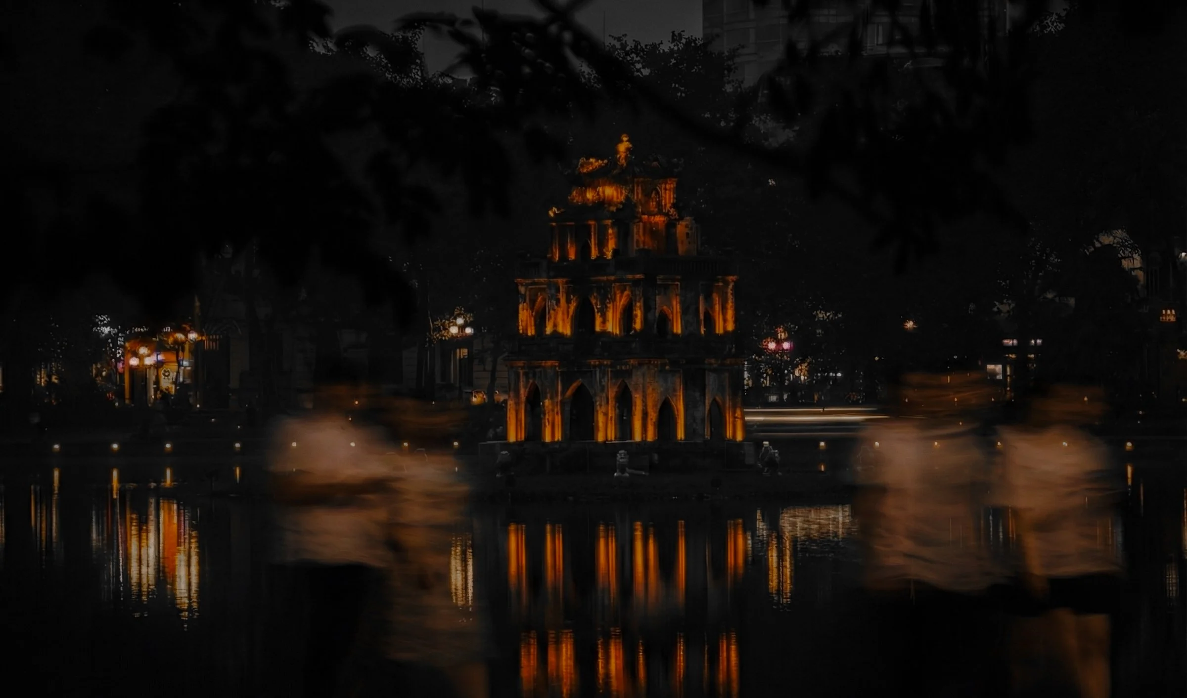 A street scene at sunset with a woman carrying baskets of produce, walking across a wet crosswalk. The street is busy with numerous motorcyclists riding in both directions, and the buildings on either side are lined with parked motorcycles. The sky displays a vibrant orange and yellow hue, contrasting with the dark cityscape.