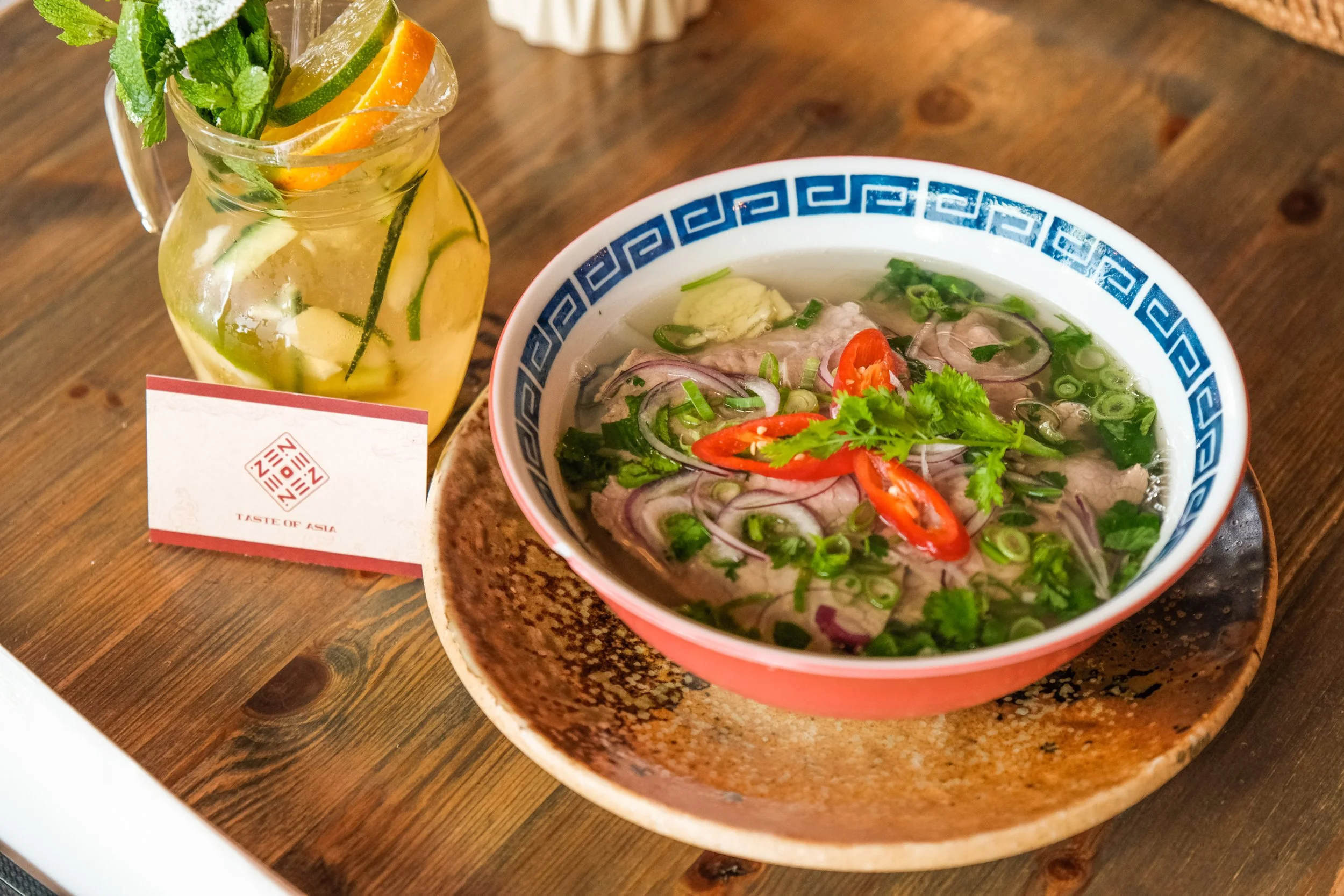 A bowl of Vietnamese pho with sliced beef, red chili peppers, green onions, and cilantro, placed on a textured brown plate. Next to it is a glass pitcher of infused water with lemon, cucumber, mint, and orange slices, accompanied by a small card that