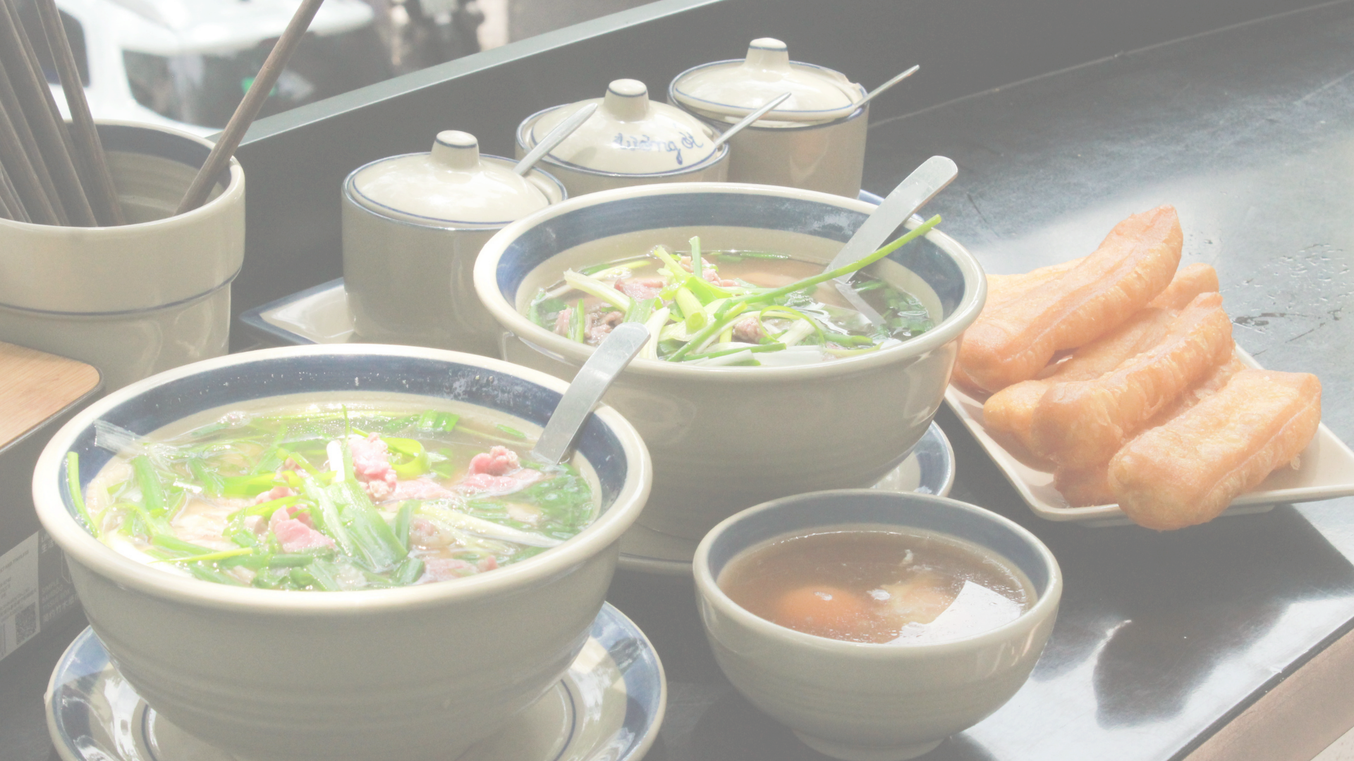 Bowl of noodle soup with green onions, slices of meat, and broth, alongside a plate of fried breadsticks, on a black table with small condiment jars in the background.