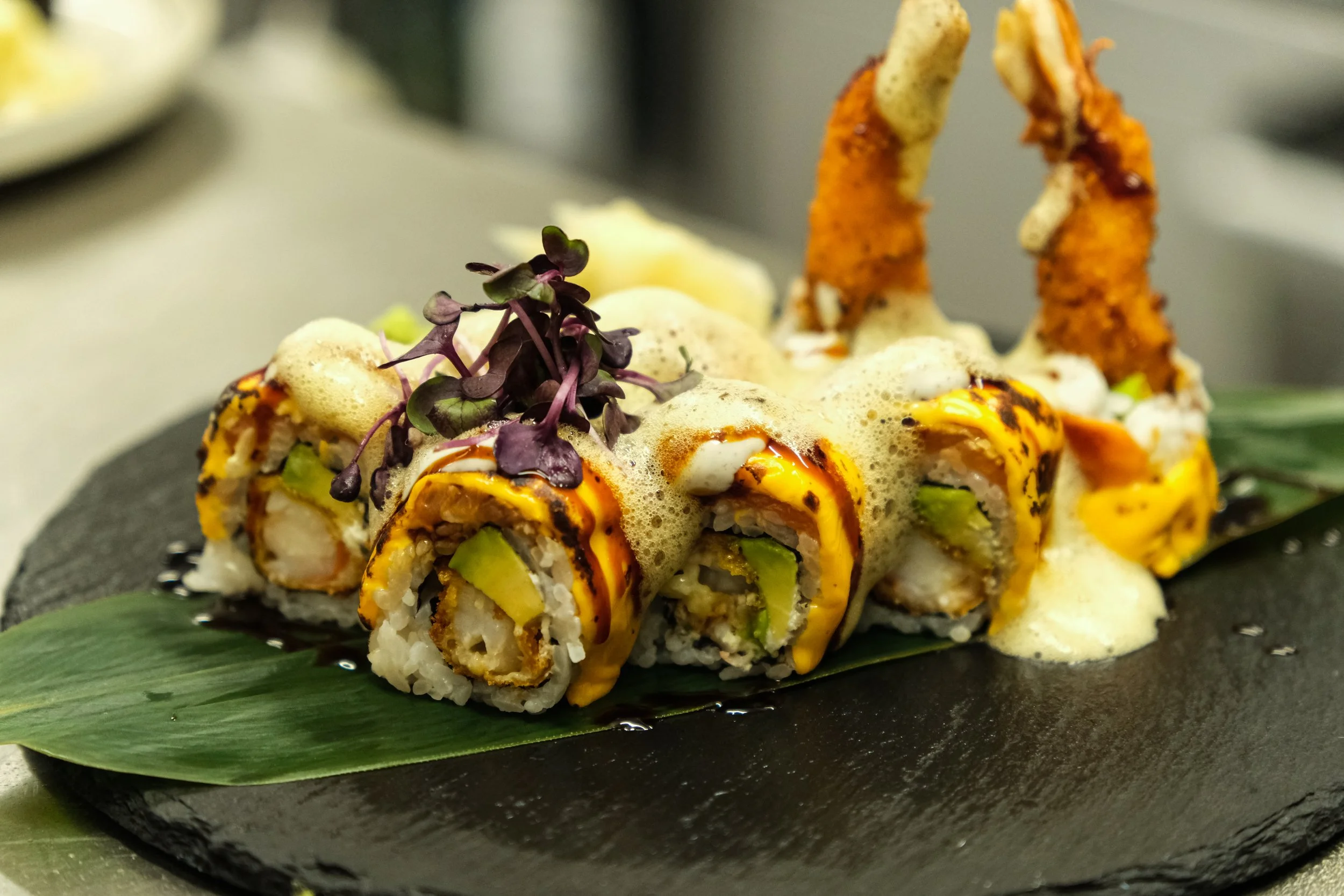 A serving of sushi rolls topped with spicy orange sauce, garnished with microgreens, served on a black slate plate with a green leaf underneath.