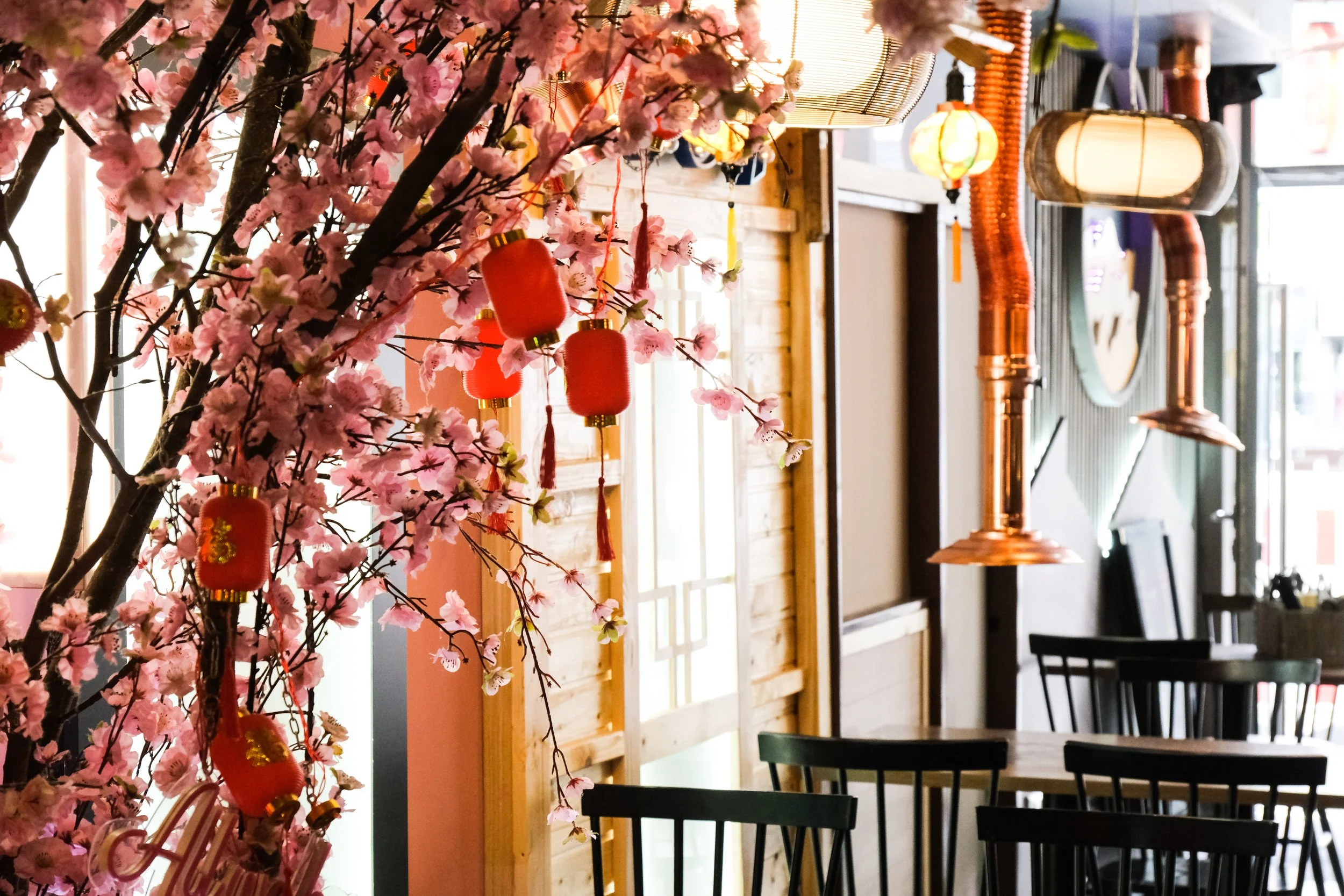 Interior of a restaurant decorated with pink cherry blossom branches and red lanterns, with tables and black chairs visible.