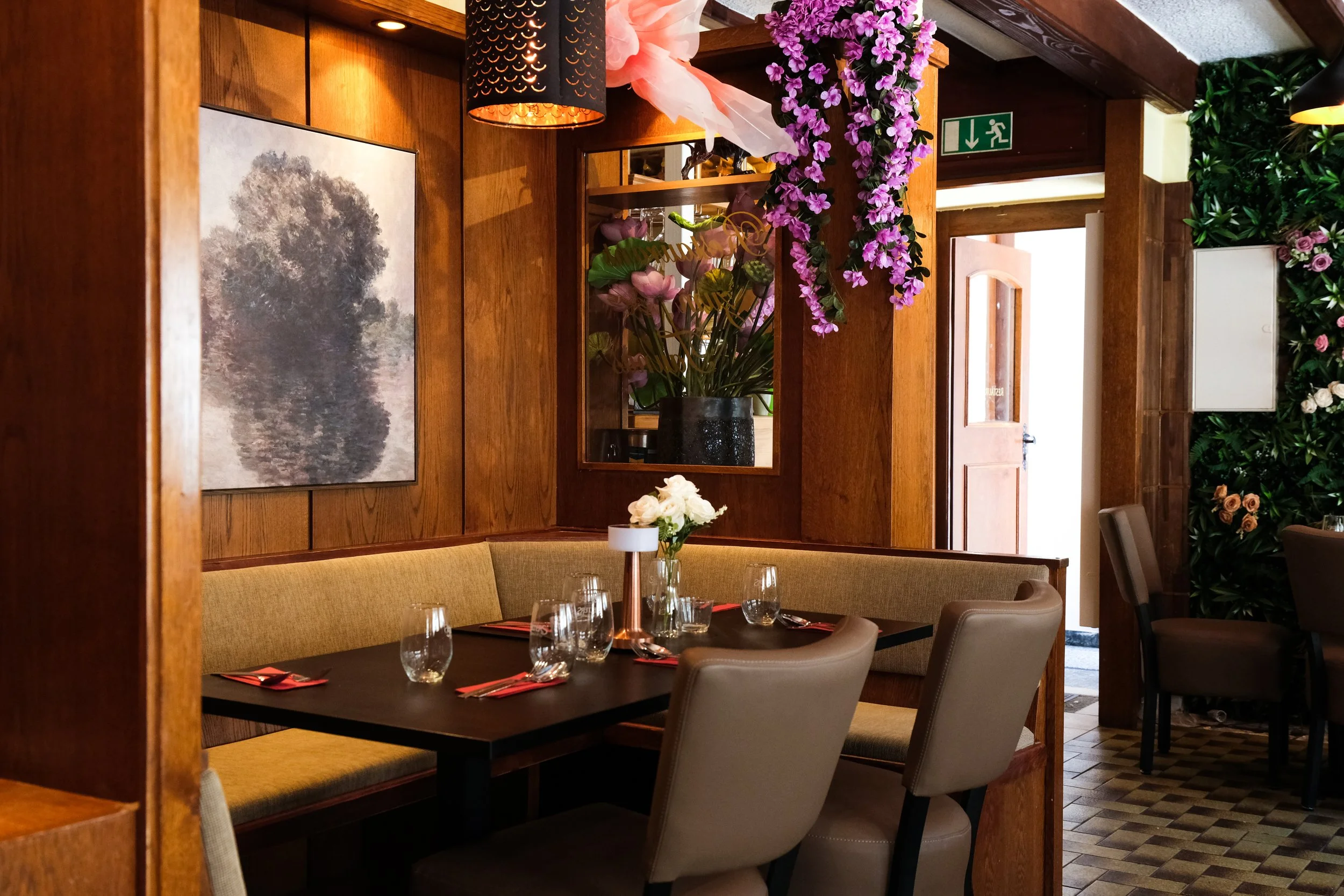 A cozy restaurant interior with a black table set with wine glasses, napkins, and a white floral centerpiece. The walls are wood-paneled with a framed black-and-white tree photograph. There are flowers hanging from the ceiling and a large potted plan