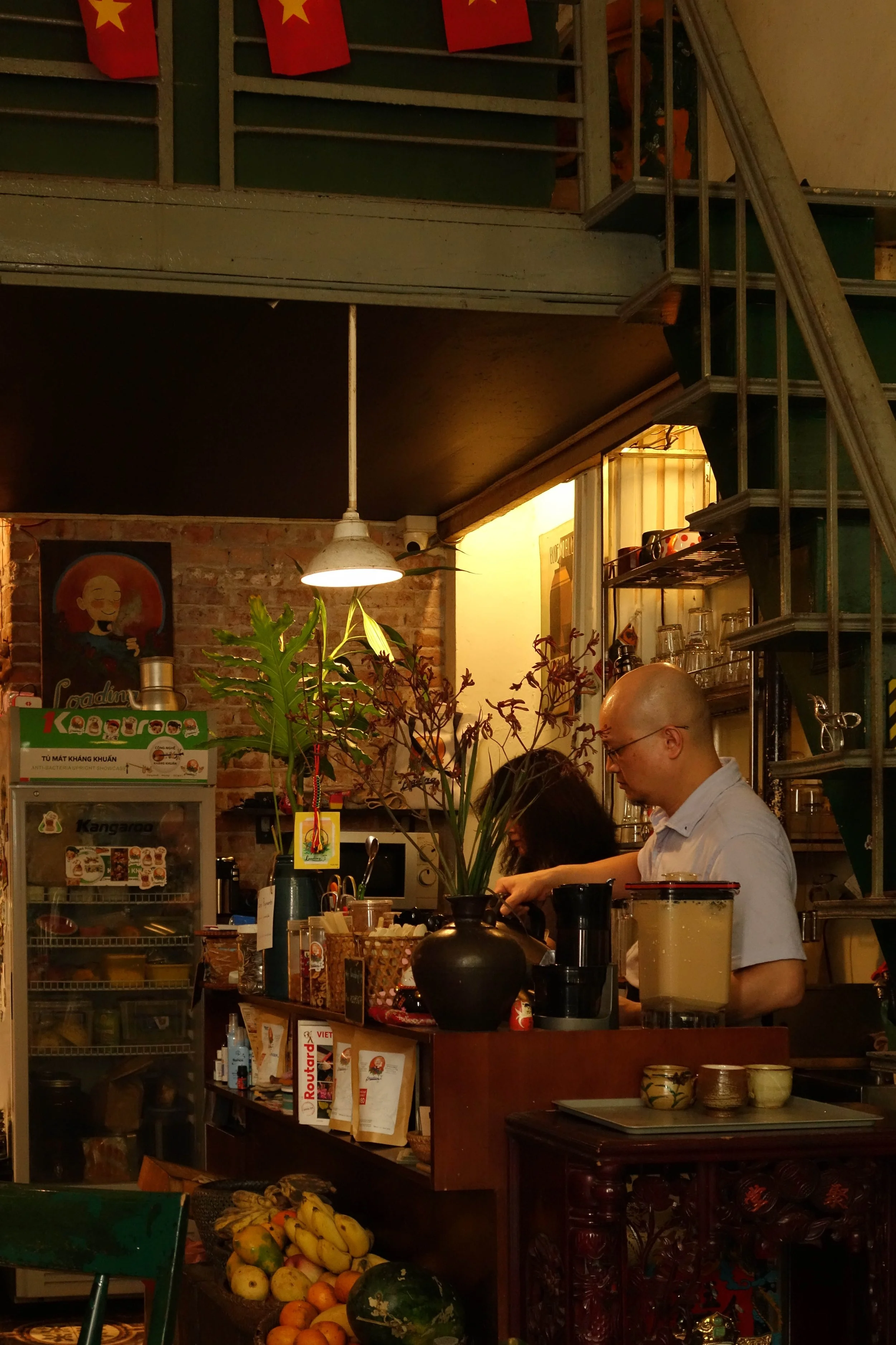 Inside a cozy cafe with brick walls and warm lighting. There are two people working behind the counter, one man with glasses and a woman, possibly baristas. The counter has various items including a black vase, a glass jar of what appears to be lemonade, and a basket of bananas, apples, and a watermelon in the foreground. Shelves with glassware and a small fridge are visible, along with a poster on the wall featuring traditional artwork and flags hanging from the ceiling.