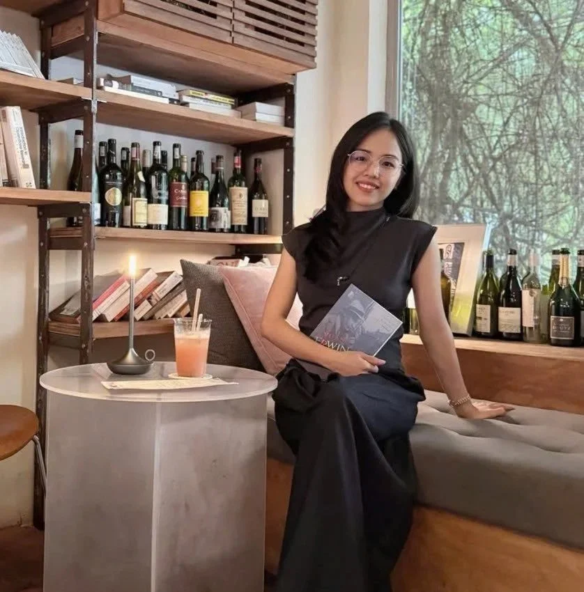 A woman sitting on a sofa in a cozy room with wooden shelves filled with wine bottles and books, holding a book, and a small round table with a lit candle and a pink drink in a glass.