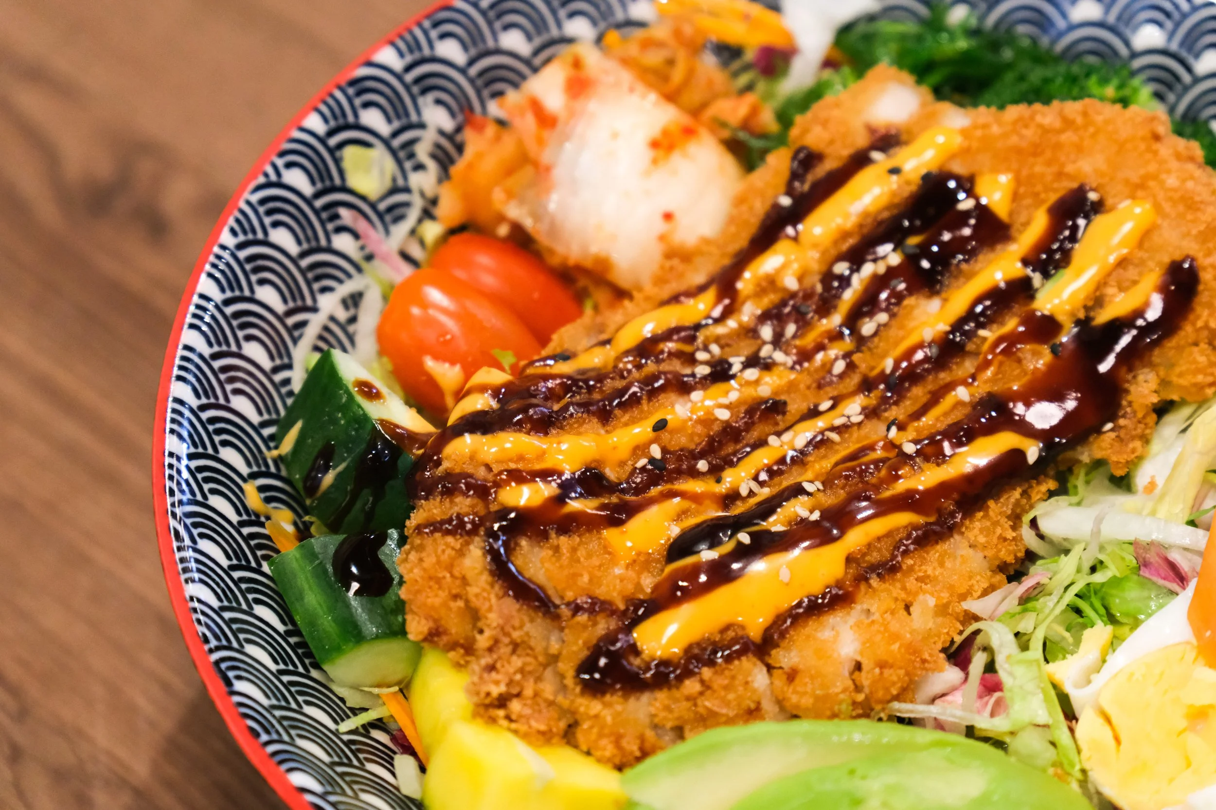 A close-up of a Japanese-style seafood and vegetable salad with fried fish fillet drizzled with black and yellow sauces, topped with sesame seeds, served in a decorative bowl.