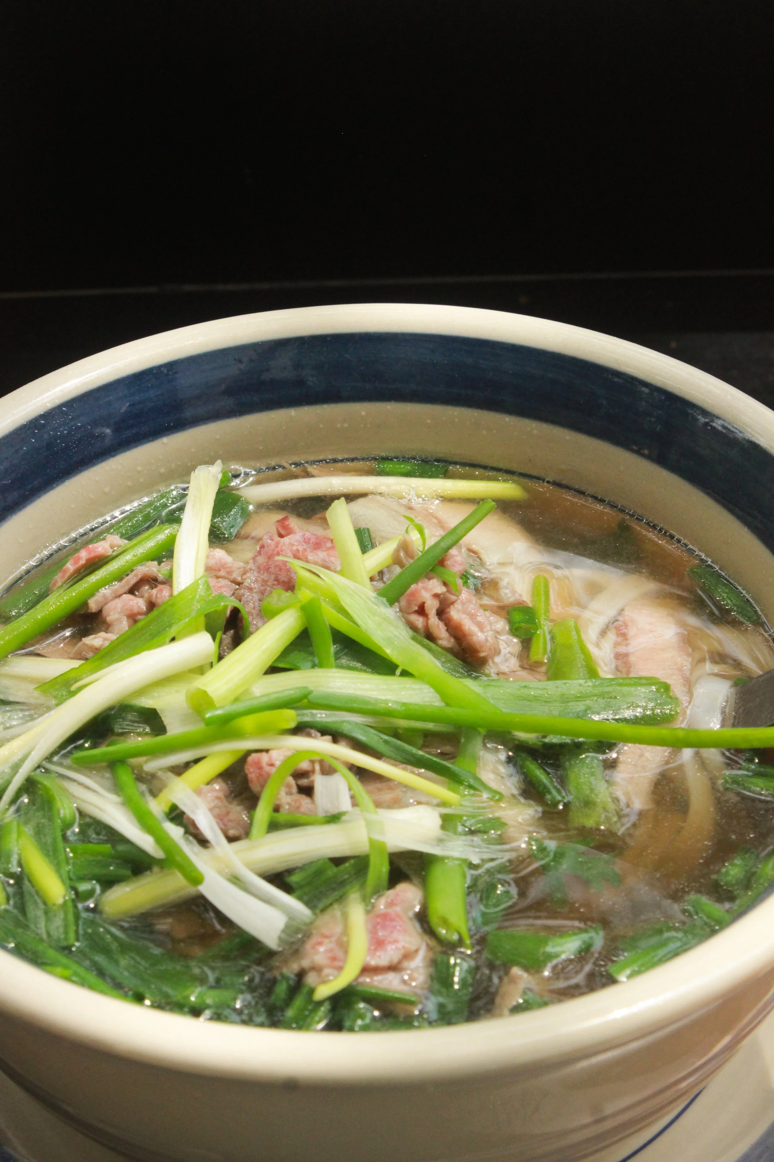 A bowl of Asian soup with sliced beef, green onions, and other vegetables in a light broth.