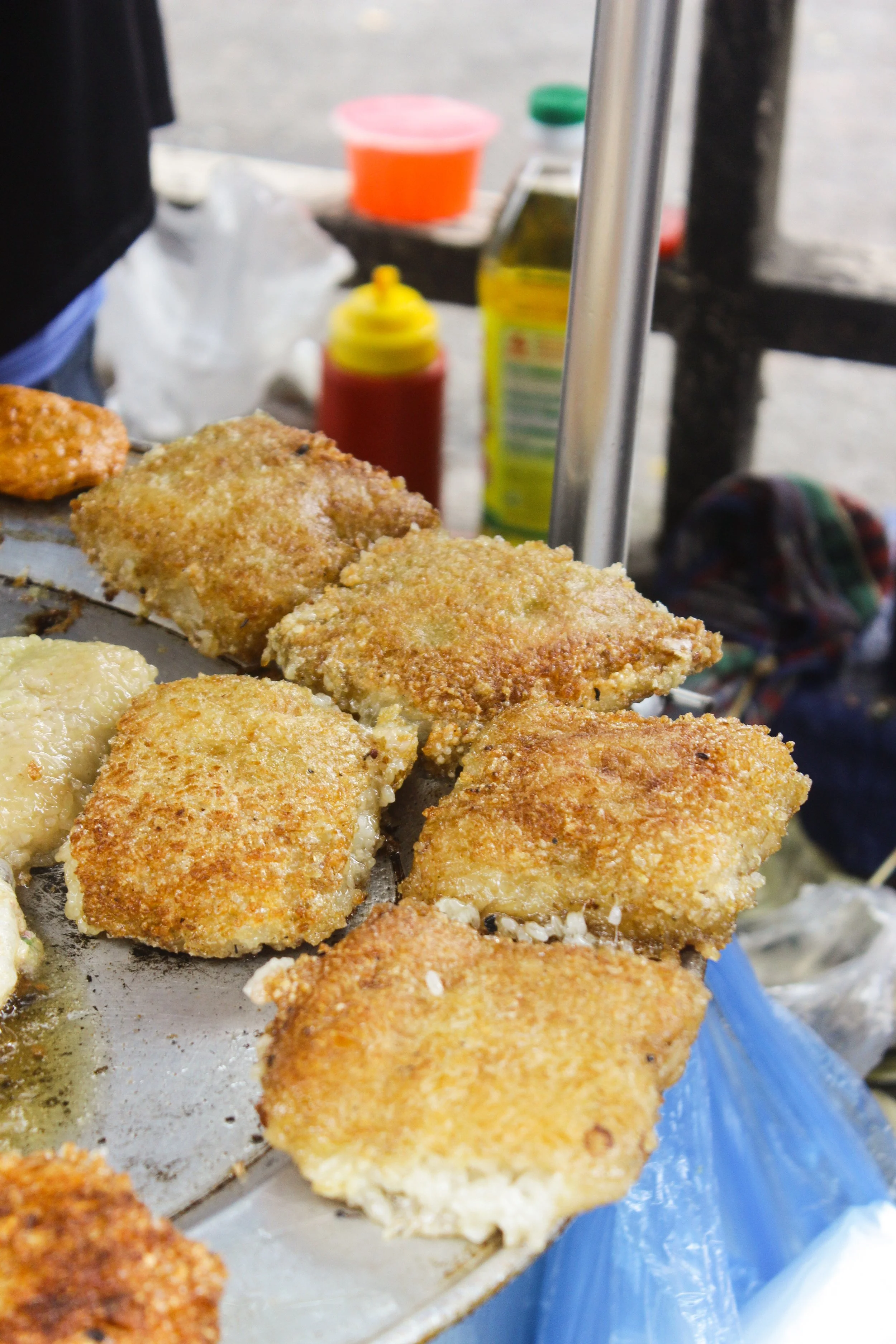 Close-up of fried food pieces, likely fish filets, on a tray at a street food stand, with condiments and bottles in the background.
