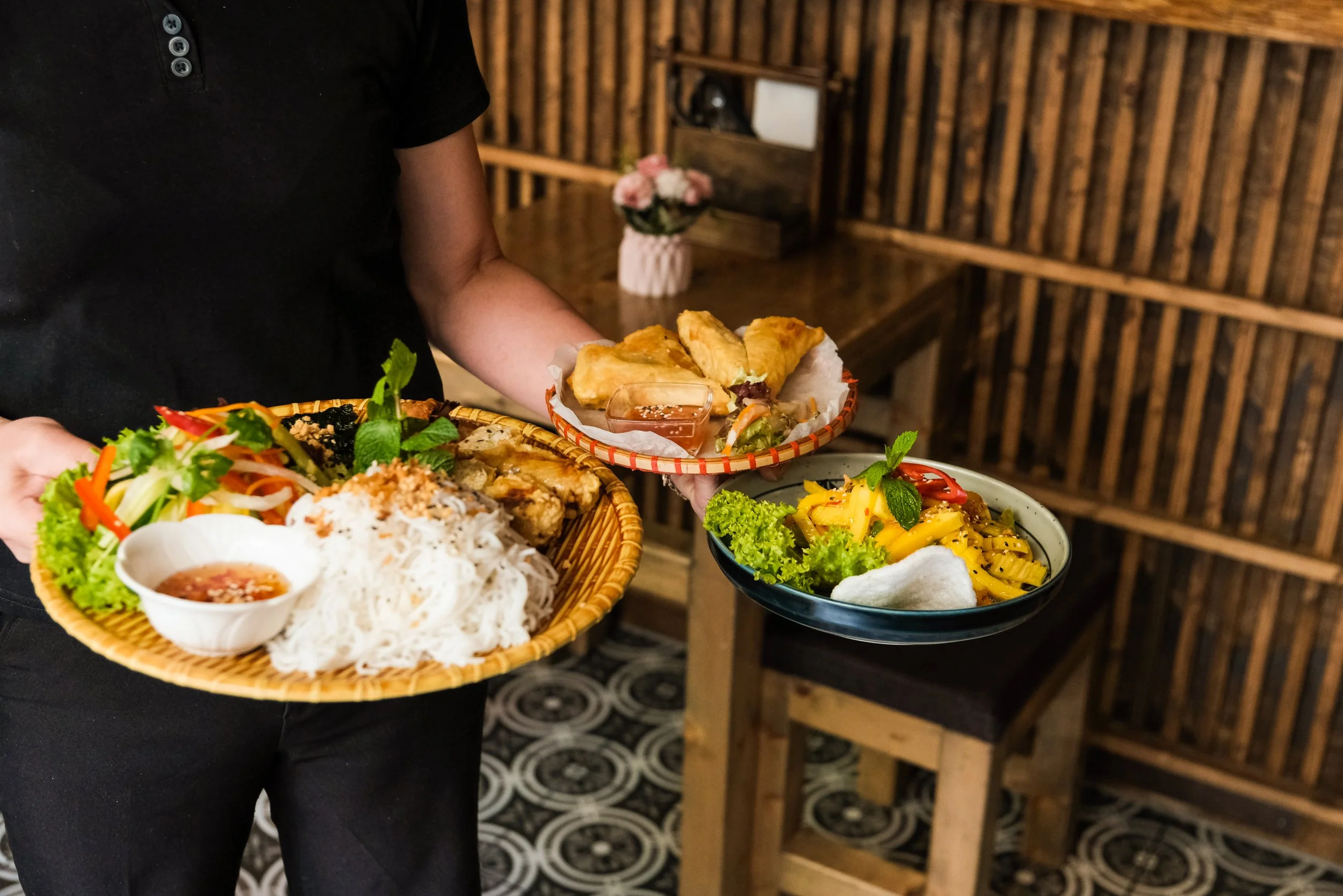 A person holding three plates of Asian cuisine in a restaurant with wooden walls and patterned floor tiles.