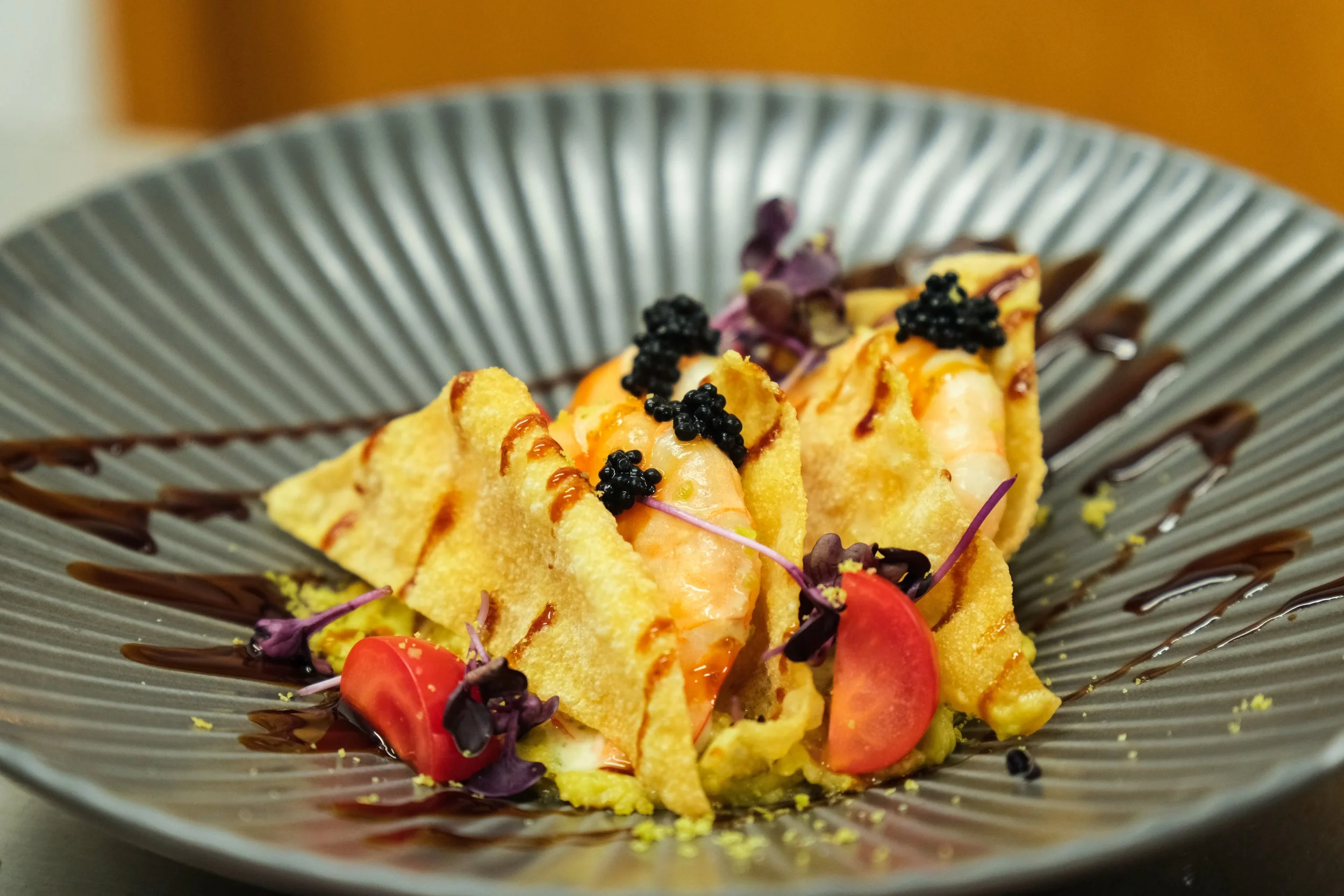 A plated dessert featuring crispy pastry filled with a creamy filling, garnished with black caviar, cherry tomato slices, microgreens, and drizzled with chocolate sauce on a decorative gray plate.