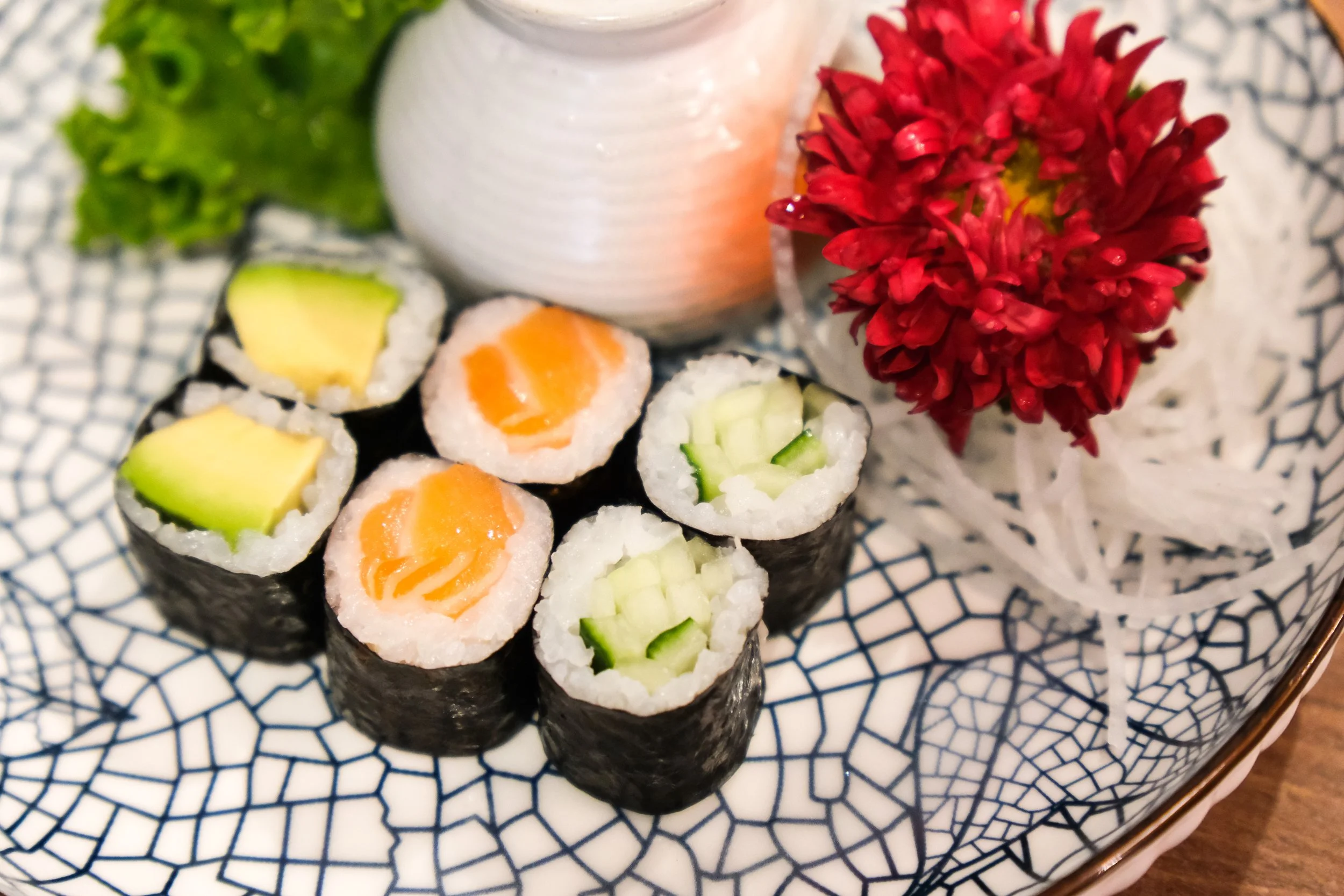 Assorted sushi rolls with cucumber, avocado, and salmon, garnished with a red flower and a white vase on a decorative plate.