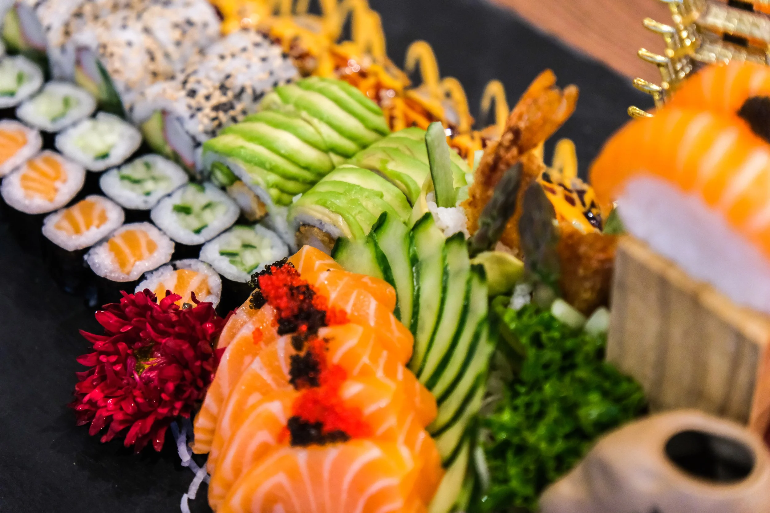 Close-up of assorted sushi rolls, including salmon, cucumber, avocado, and tempura, arranged on a black platter with garnishes and flowers.