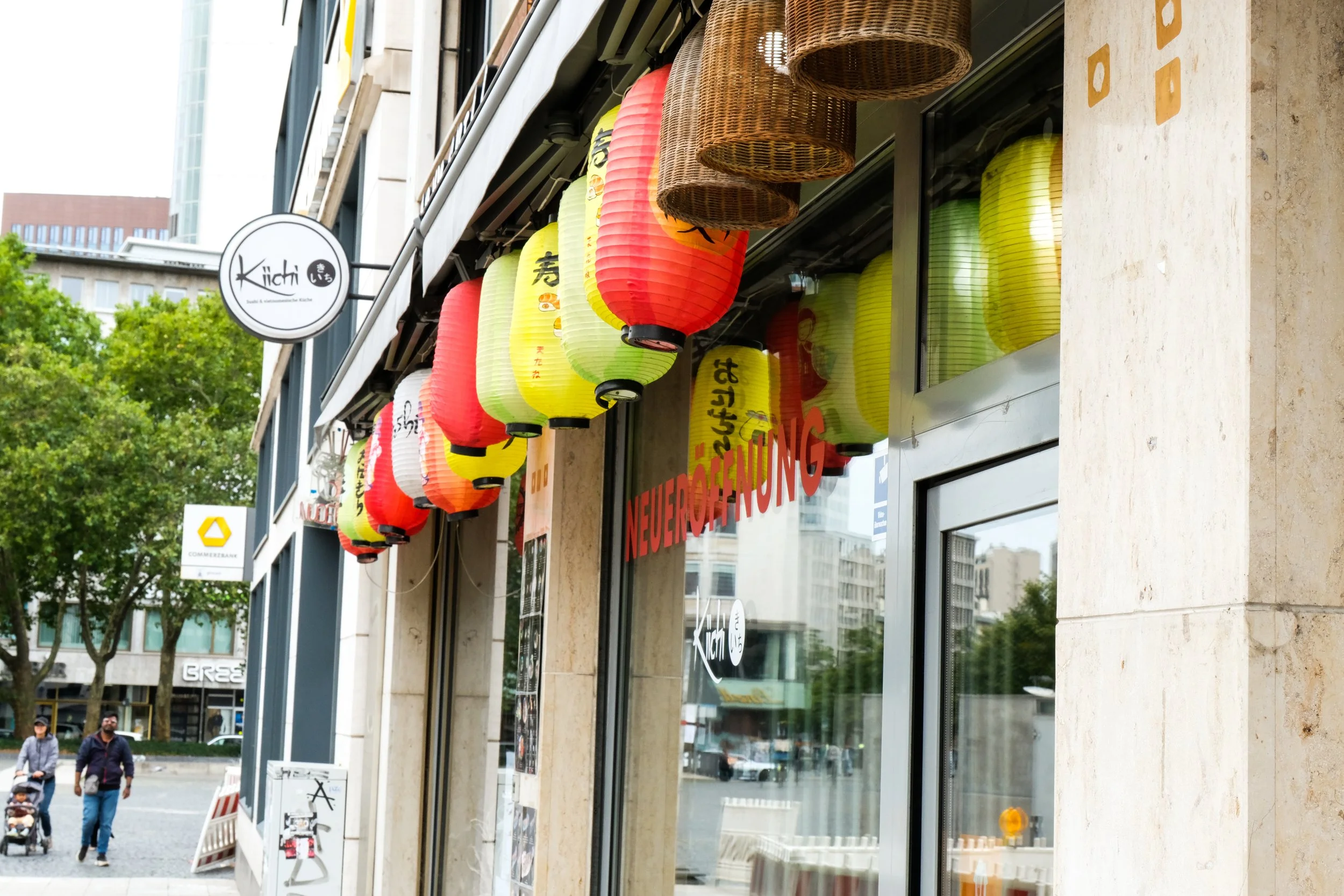 Exterior of a restaurant or shop with colorful Japanese paper lanterns hanging above the entrance, including red, yellow, and green lanterns with Japanese characters. Signs indicate it is newly opened.