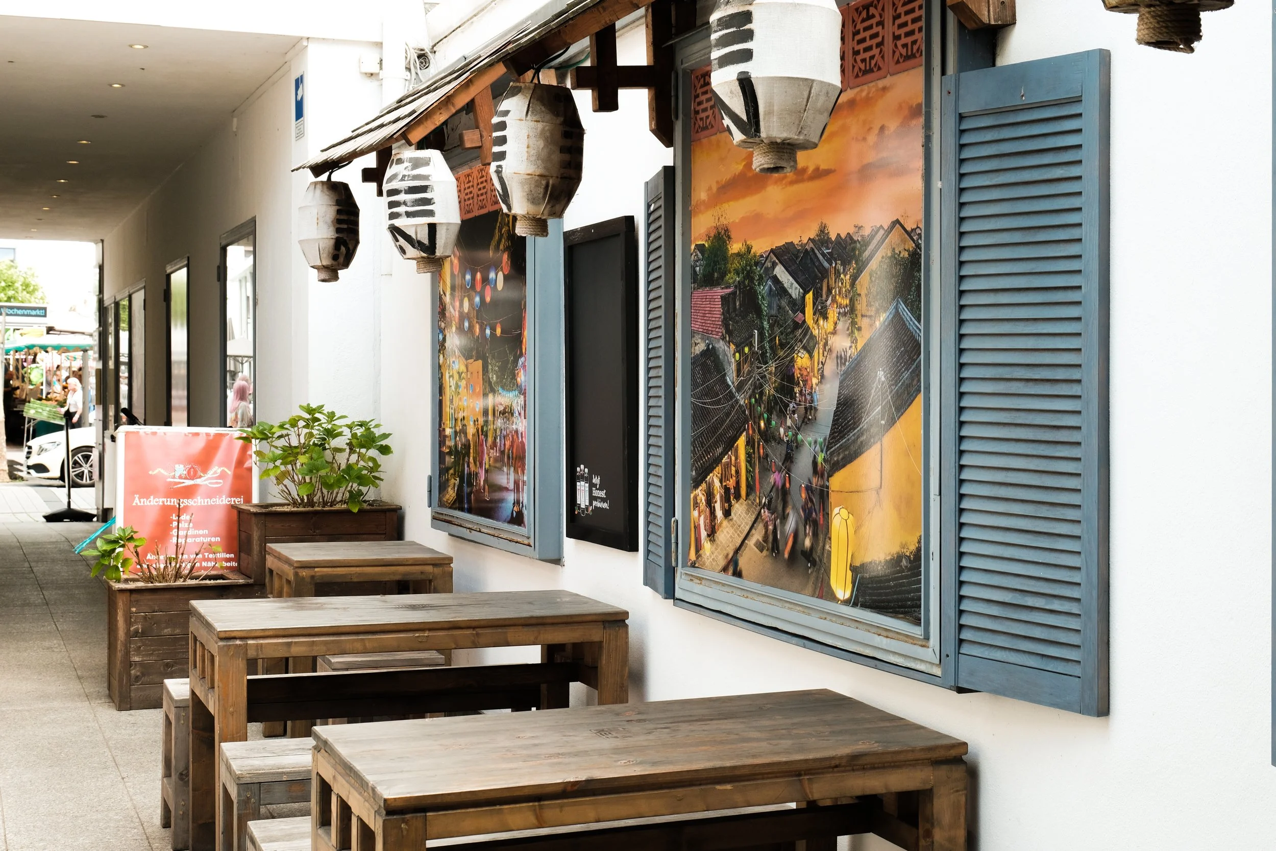 Outdoor seating area with wooden tables and potted plants, decorated with hanging lanterns, outside a building with large colorful street scene paintings on the wall.