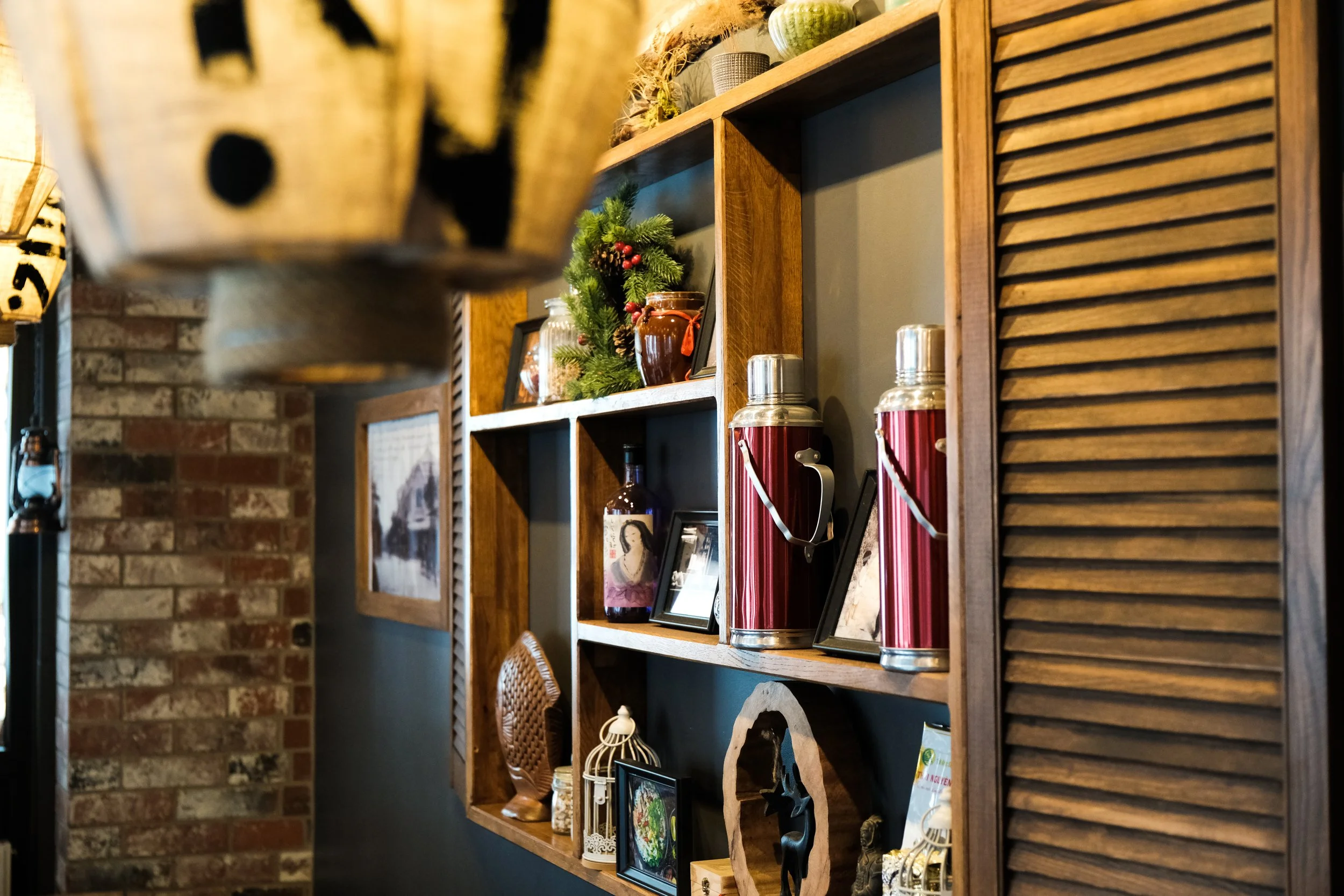 Wooden shelving unit with various decorations, including framed photos, vases, Christmas decorations, and red thermoses, against a wall with brick and painted texture.