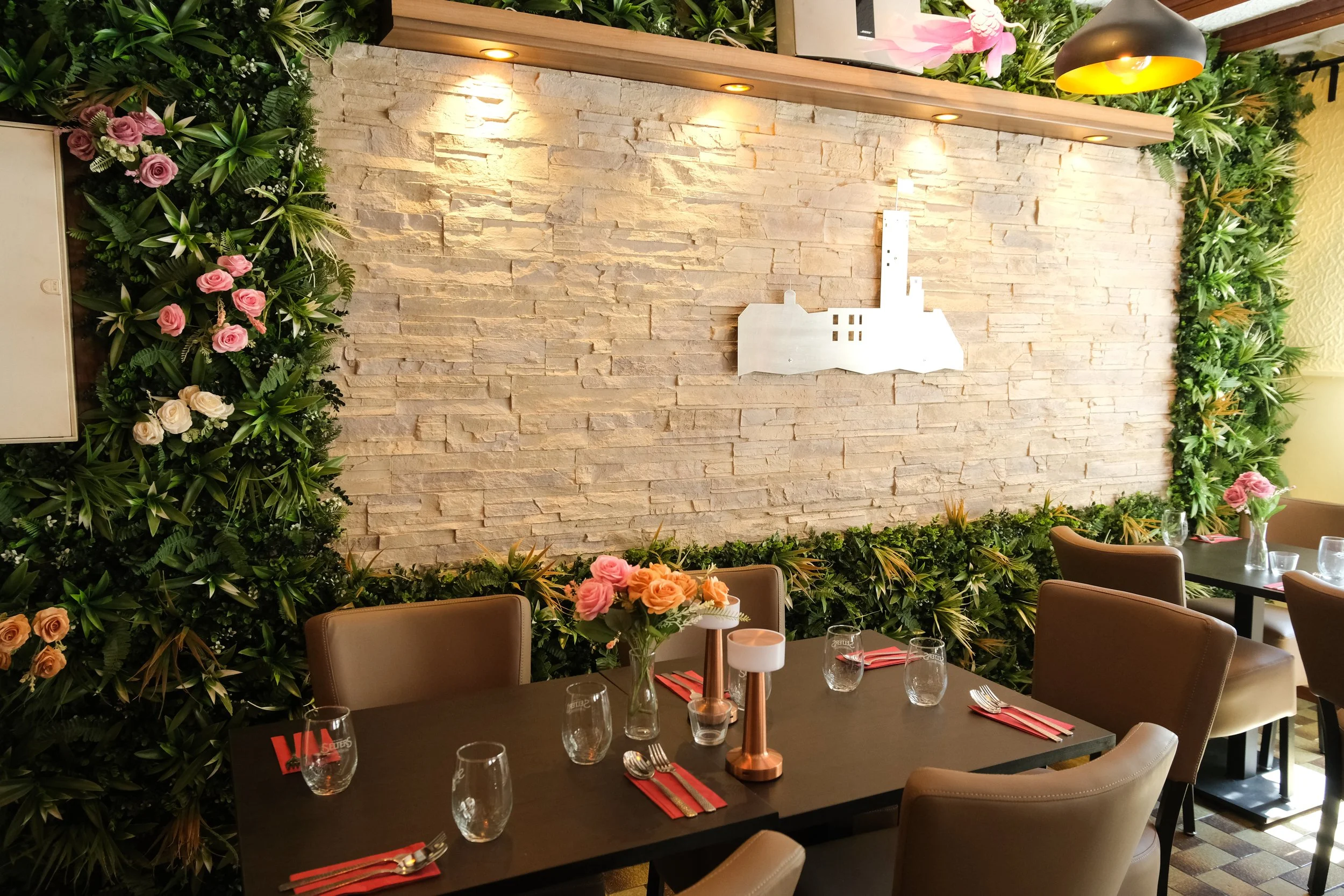 A dining area with a black table, brown chairs, and a floral centerpiece. The wall behind is decorated with white stone, greenery, and pink and white flowers. Silverware and glasses are set on the table.