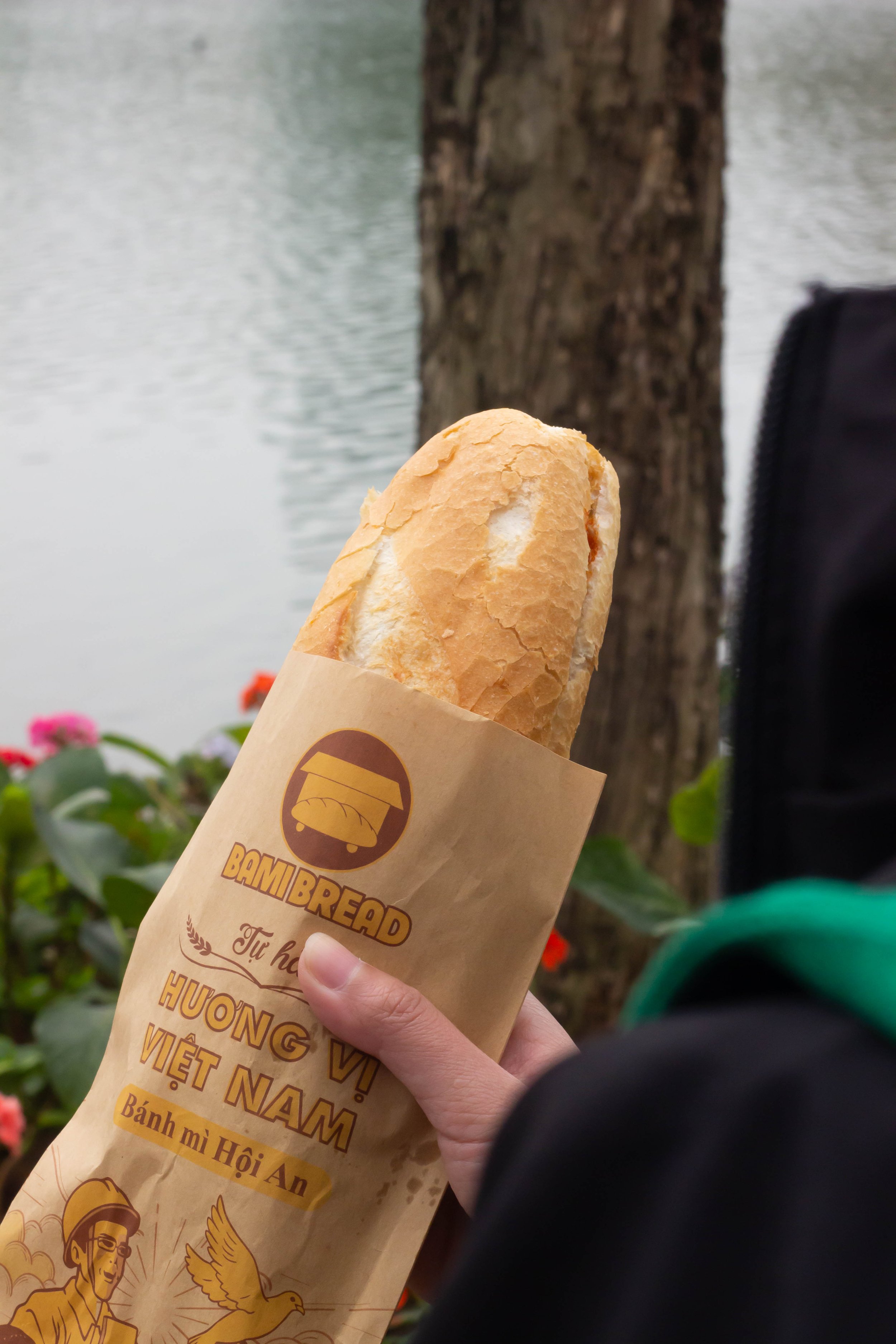A person holding a baguette sandwich wrapped in paper with the logo and text 'Bami Bread' against a backdrop of a tree and water.
