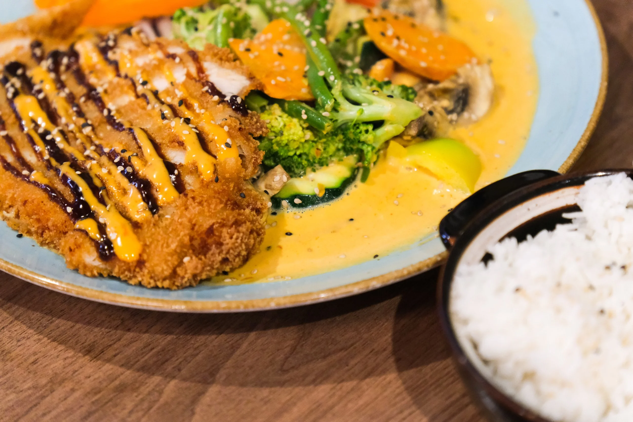 Crispy fried chicken cutlet with drizzles of mustard and barbecue sauce served with broccoli, carrots, and zucchini in a creamy yellow curry sauce, alongside a bowl of white rice.