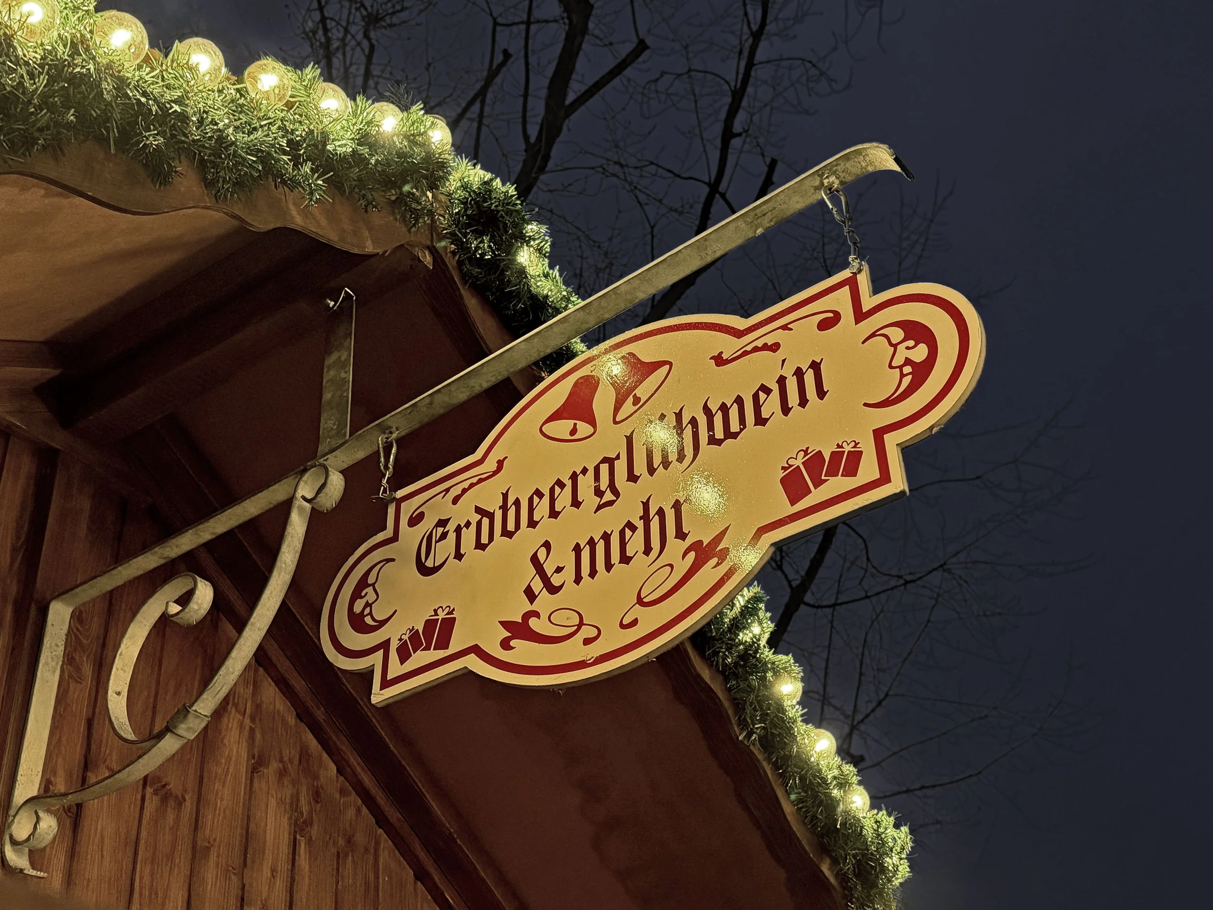 Geschild Metallzeichen mit Weihnachtsschmuck und Lichterkette, das die Begrüßung 'Erdbeeralarmwein & mehr' anzeigt, an einem Holzwand, im Dunkeln mit bläulichem Himmel im Hintergrund.