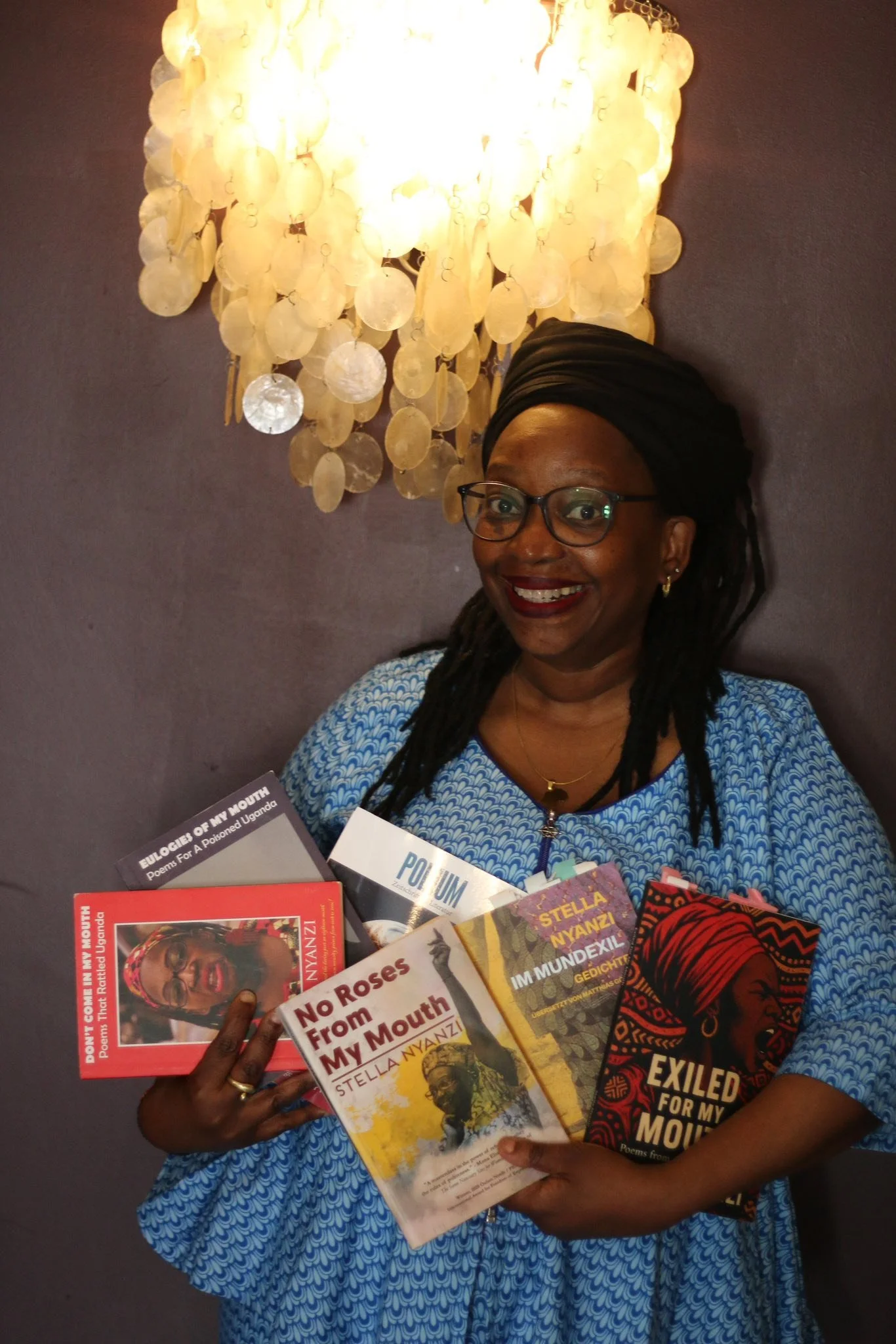 A woman smiling, wearing glasses and a black headwrap, holding several books with titles related to poetry and Uganda, standing in front of a dark wall and a chandelier made of disc-shaped shells.