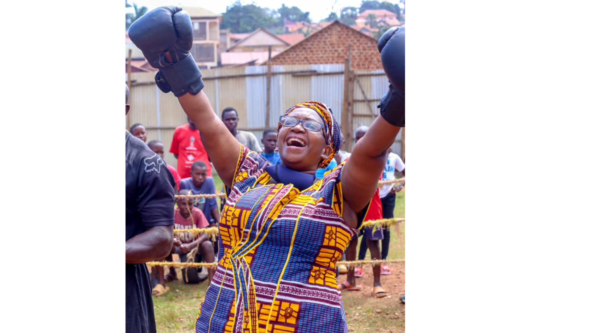 Woman in colorful traditional African clothing raising her arms in victory, surrounded by onlookers at outdoor event.