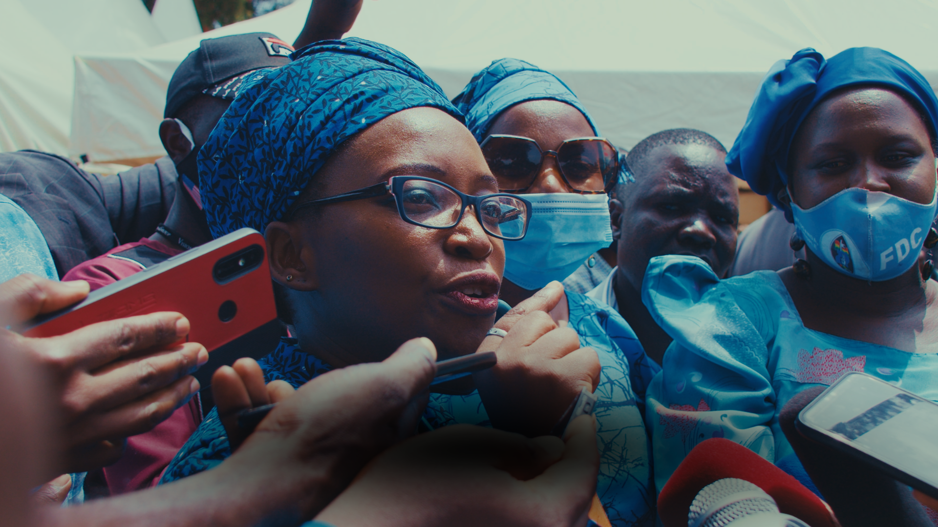 A group of people, including women wearing headscarves and face masks, gathered closely together, some holding smartphones, at an outdoor event or press conference.