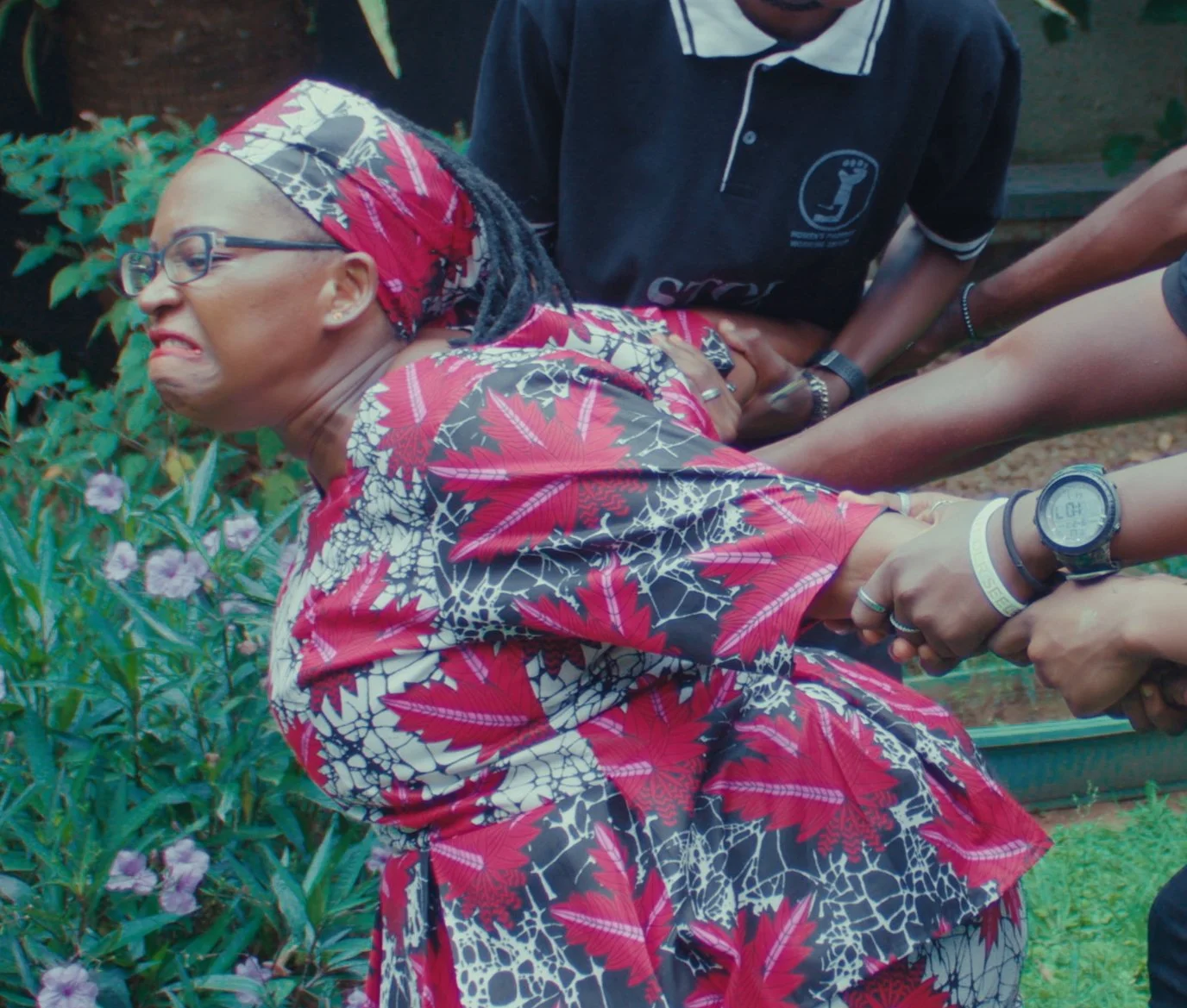 A woman with glasses and a red floral headscarf is being detained, pulling her arms behind her back by multiple people.