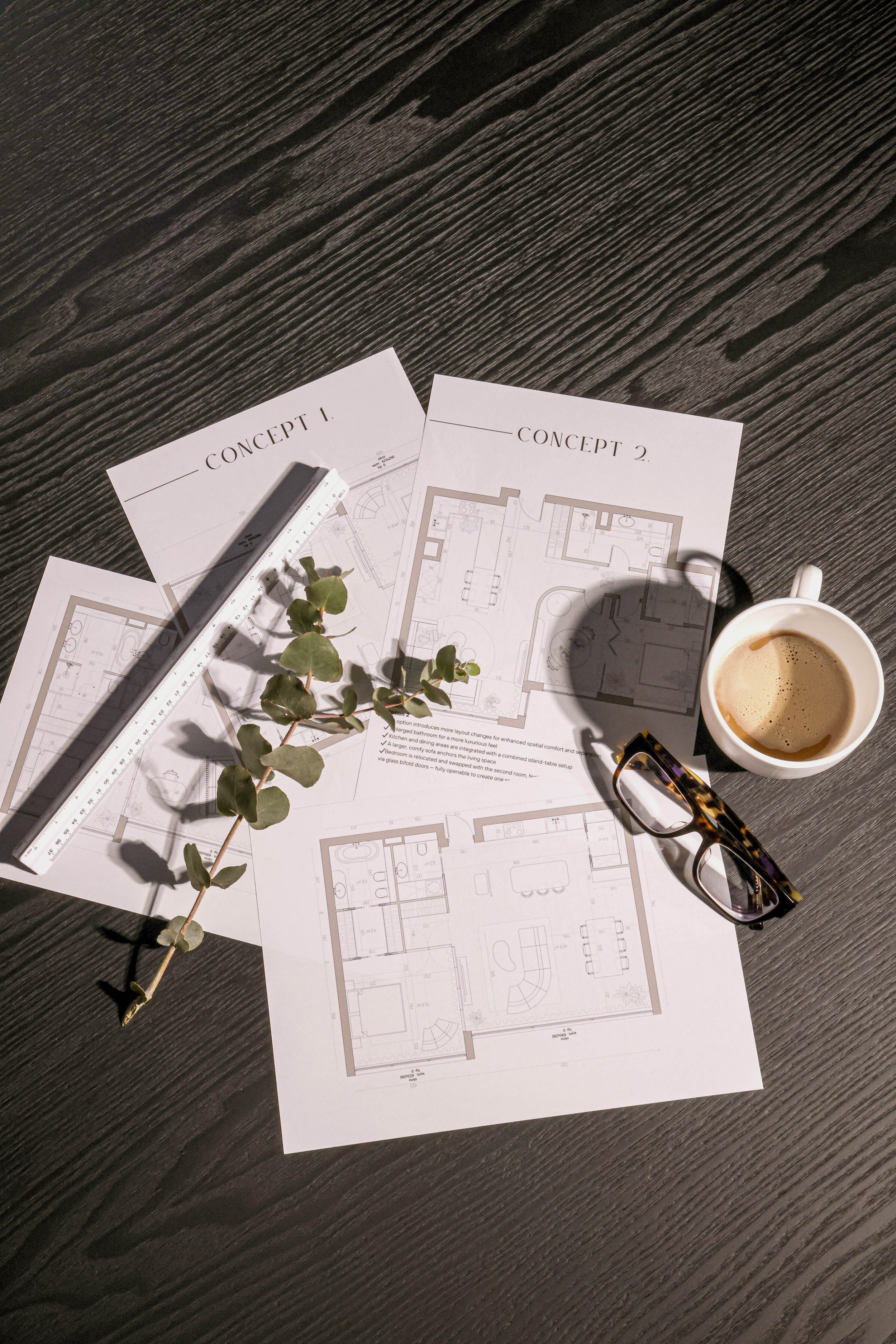 Architectural blueprints labeled 'Concept 1' and 'Concept 2' on a dark wooden table, with a metal ruler, eucalyptus branch, cup of coffee, and tortoise-shell glasses. Formalivinh, Beirut Lebanon.