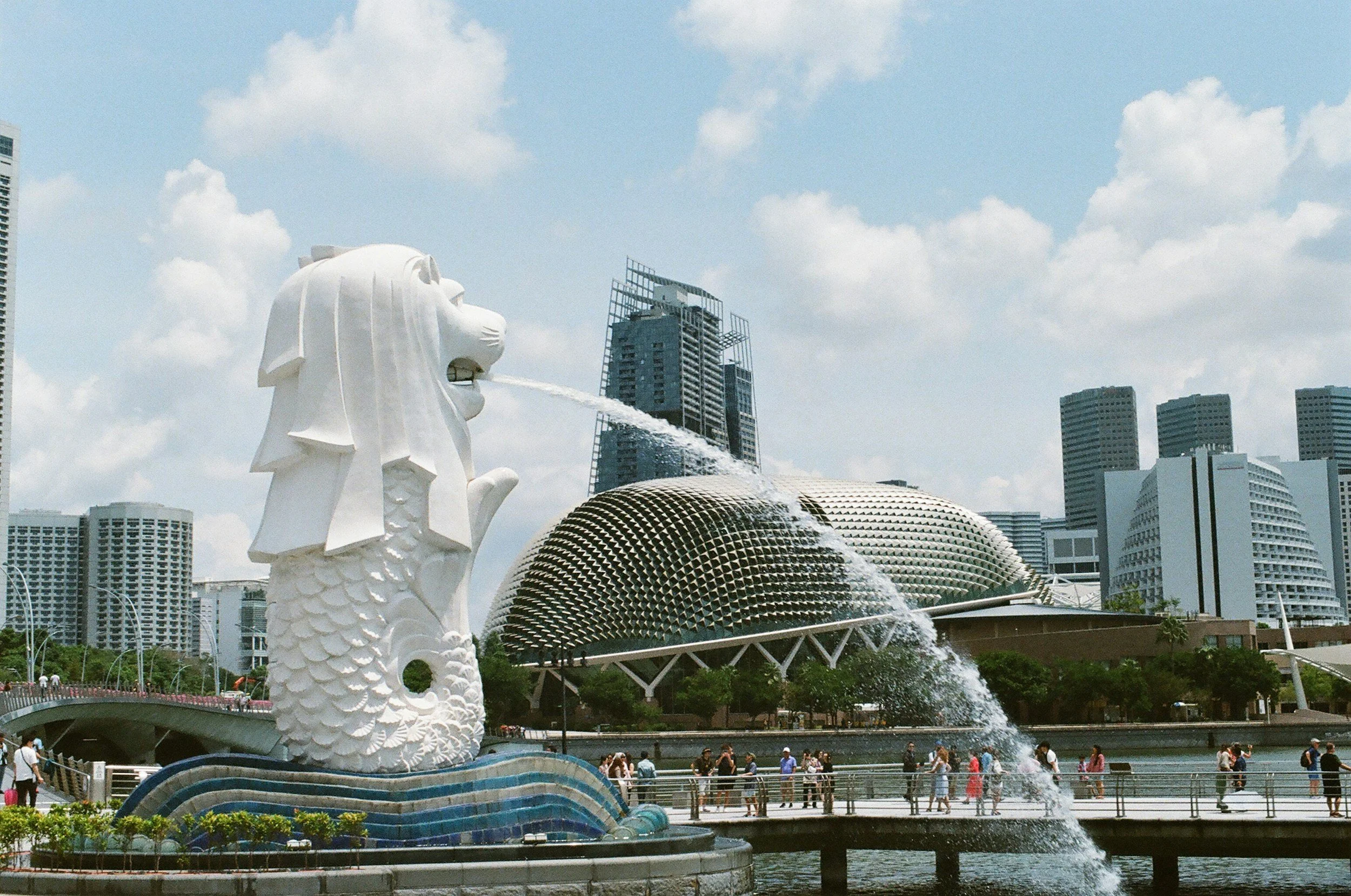 Singapore's Merlion statue with a lion head and fish body, spouting water from its mouth, located in a cityscape with modern skyscrapers and the Esplanade building in the background.