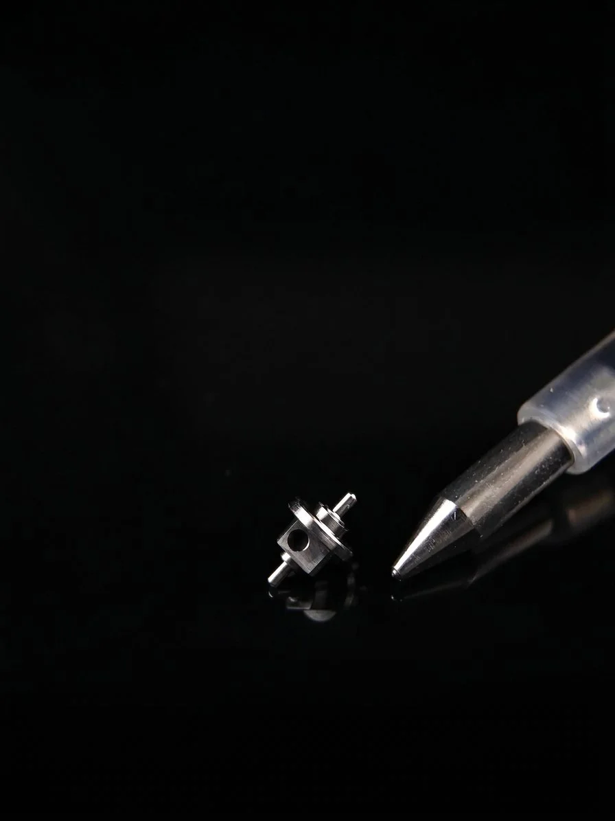 A tiny high precision metal mechanical part with holes and protrusions, next to a pen tip on a black reflective surface.