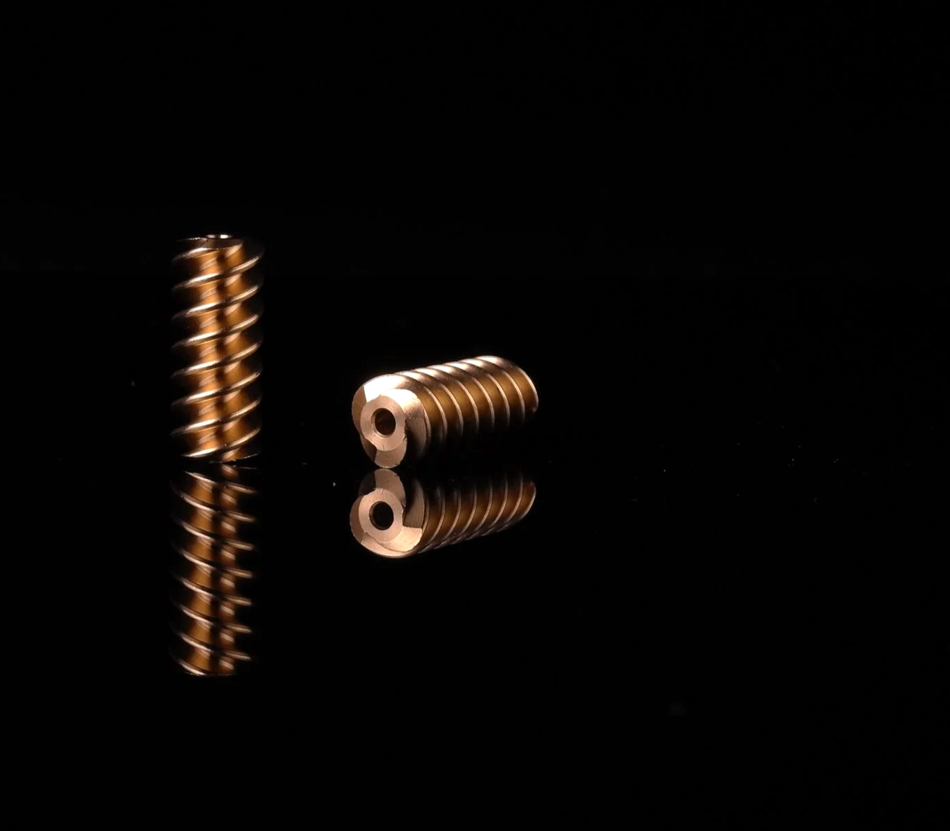 Two worm gears with a bronze finish on a reflective black surface, one standing upright and the other lying down.