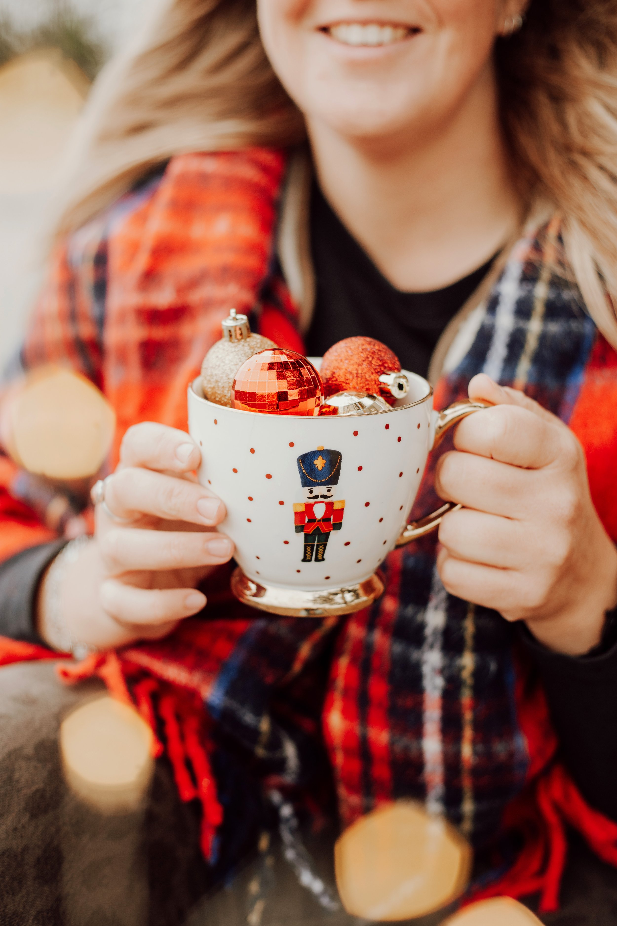 Person holding a mug filled with Christmas ornaments, wearing a plaid jacket and smiling.