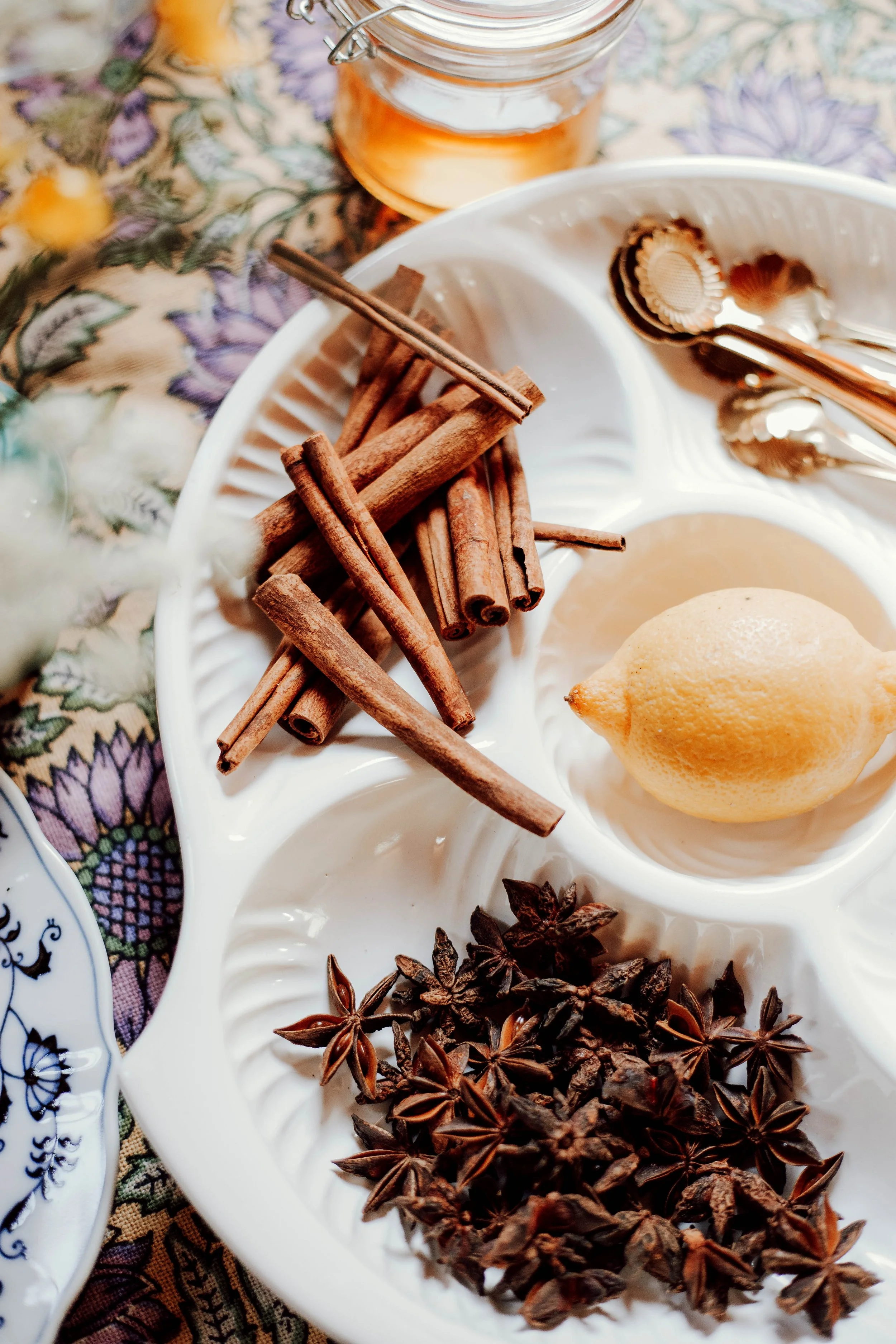 A white segmented plate containing cinnamon sticks, star anise, a lemon, and a jar of honey on a floral tablecloth.