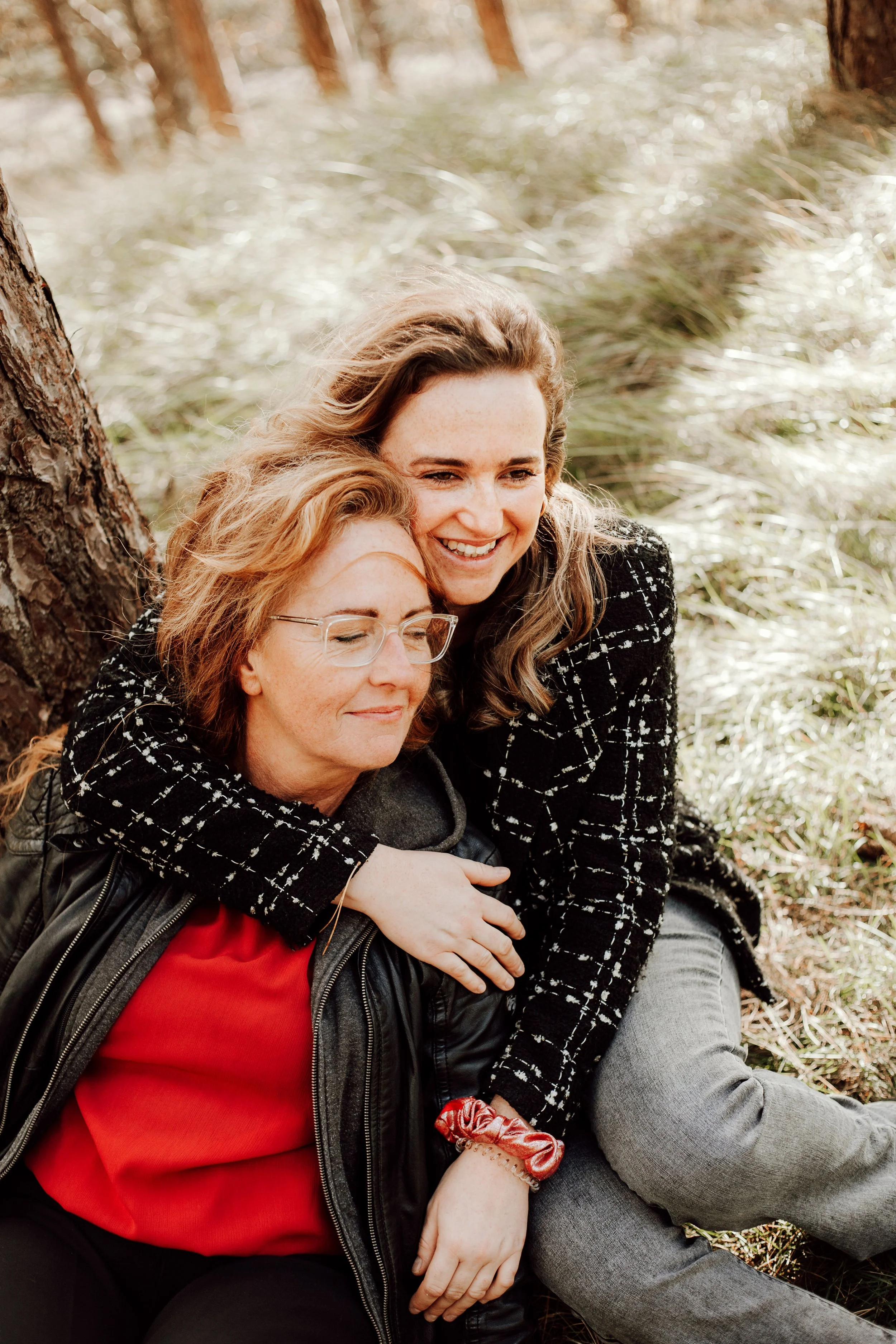 Two women sitting outdoors near a tree, hugging and smiling, with a grassy background.