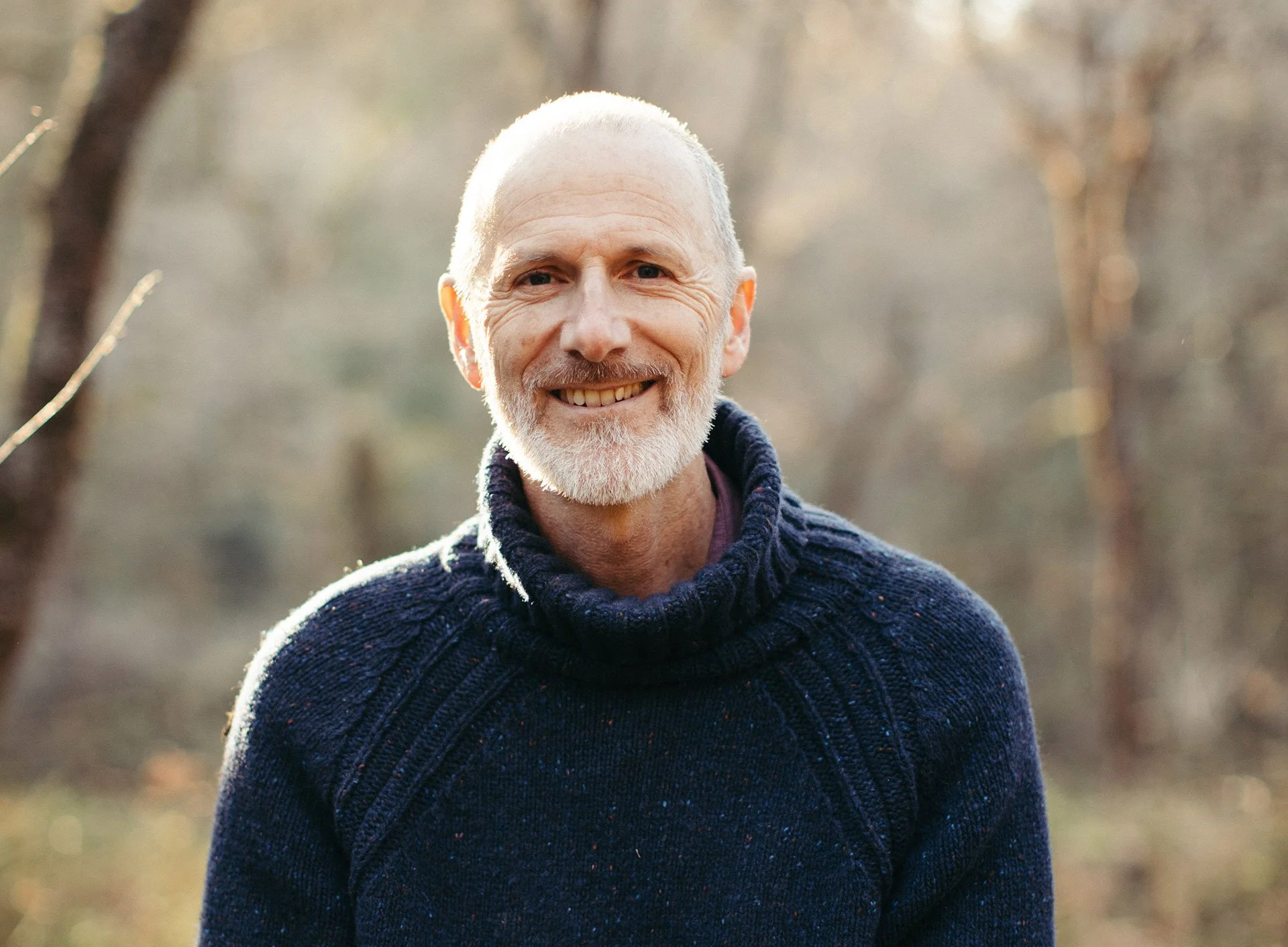 A smiling older man with a short gray beard and short gray hair outdoors during daytime, wearing a dark blue knitted sweater, with blurred trees in the background.