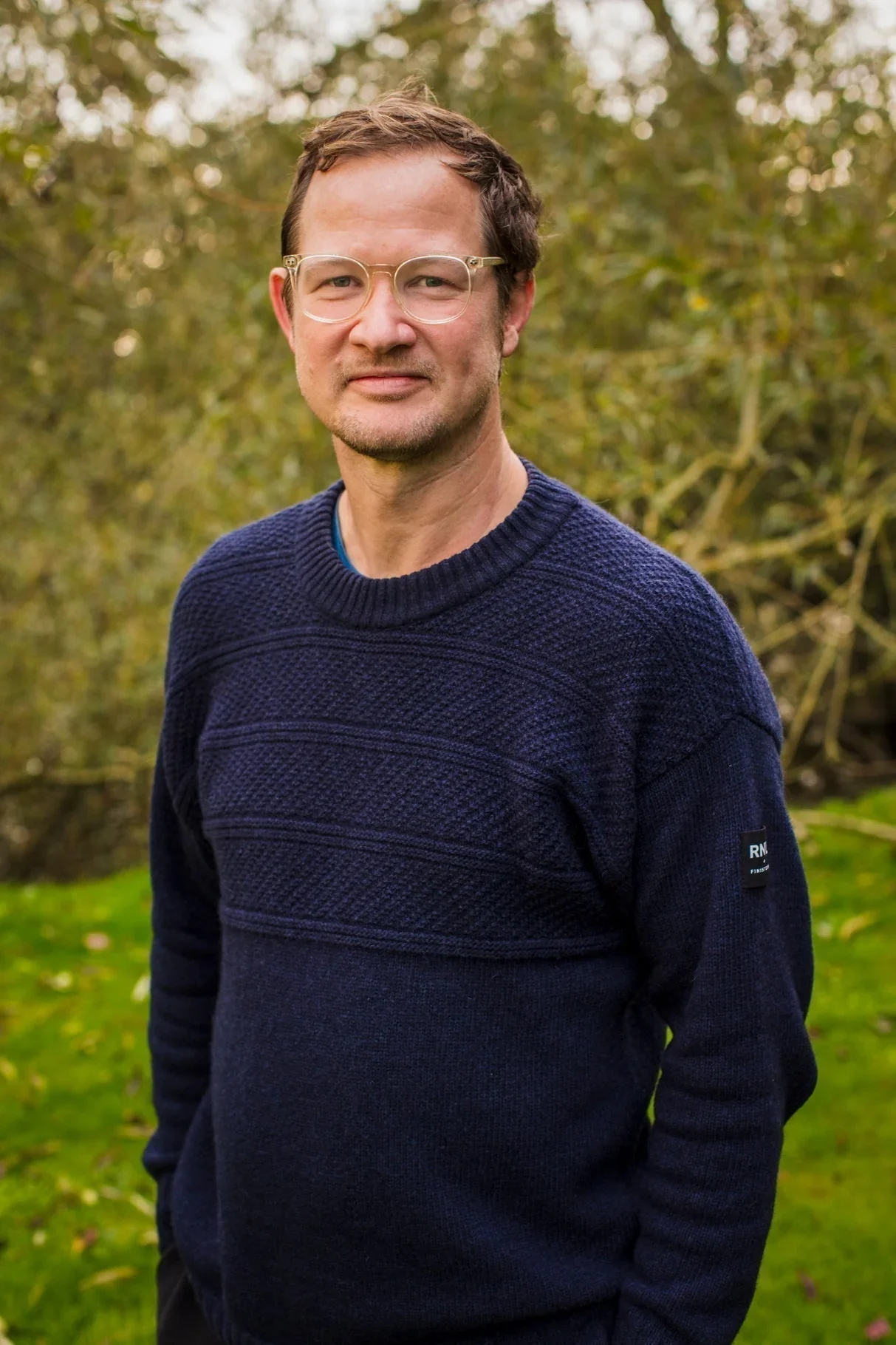 A man with glasses and short brown hair standing outdoors in a park with green and yellow foliage in the background, wearing a navy blue sweater.