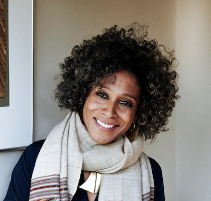 Woman with curly hair smiling, wearing a beige scarf and dark clothing, standing indoors.
