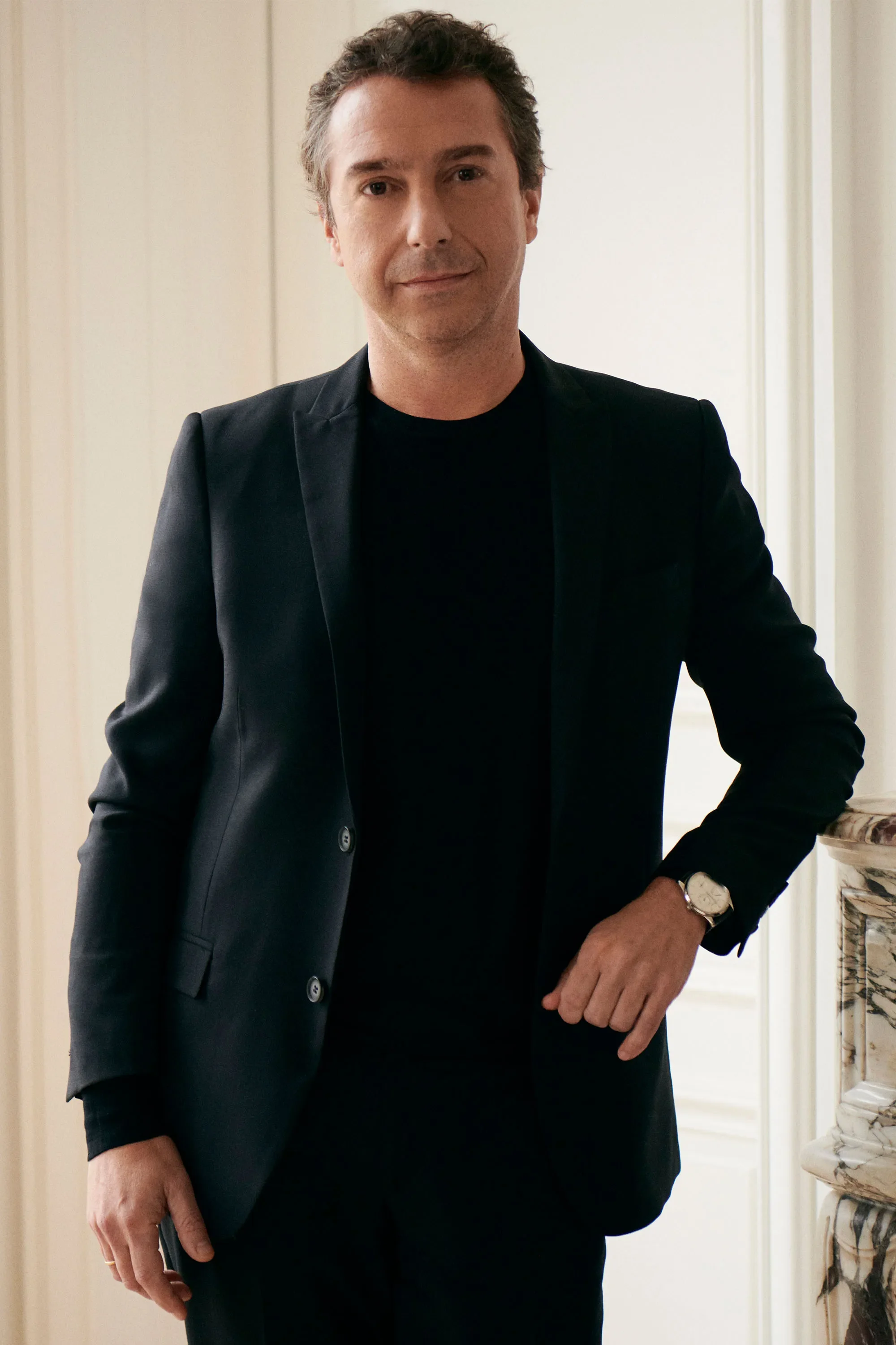 A man wearing a black blazer and black shirt, standing indoors next to a marble fireplace, with a watch on his left wrist, smiling softly.