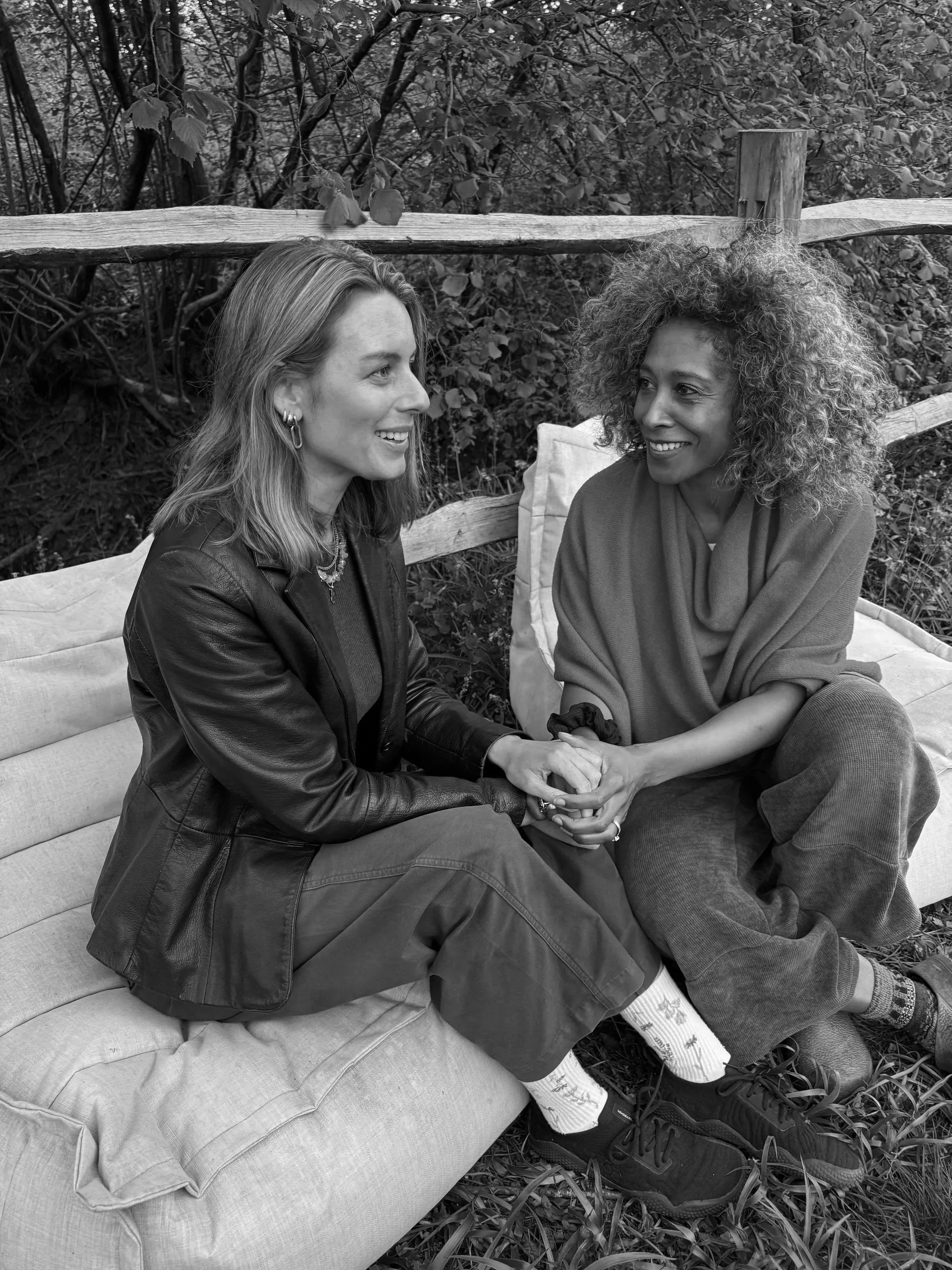 Two women sitting on a cushioned outdoor bench, holding hands and smiling at each other, surrounded by trees and foliage.