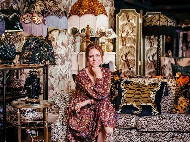 A woman sitting on a leopard print sofa in a room decorated with various lampshades, wallpaper, and decorative elements.