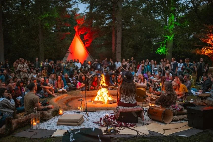 A large group of people gathered outdoors around a campfire, listening to two musicians playing guitars, with a teepee decorated with colorful lights in the background.