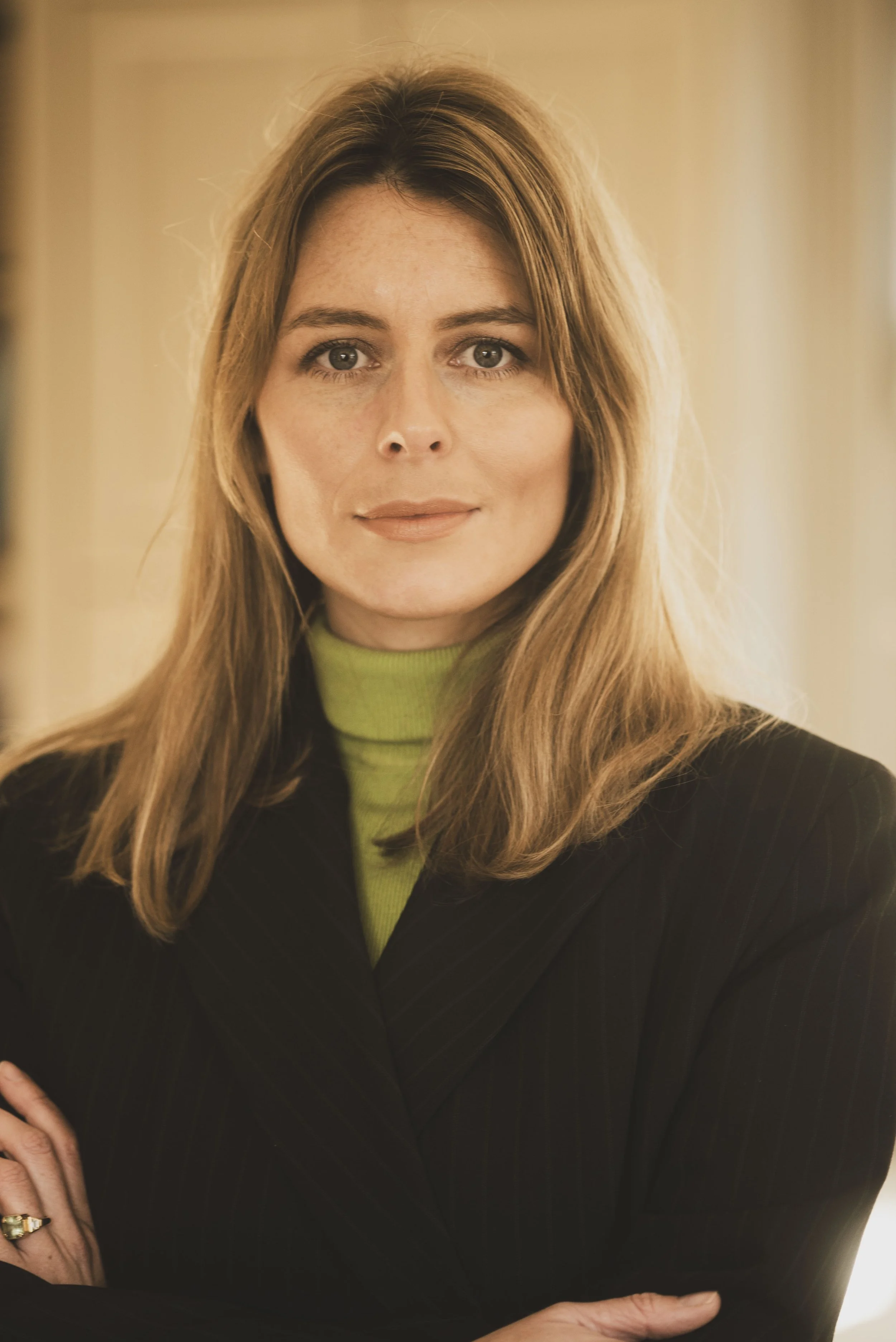 Close-up of a woman with long, light brown hair and light skin, wearing a lime green turtleneck and a dark blazer, looking directly at the camera with a slight smile.