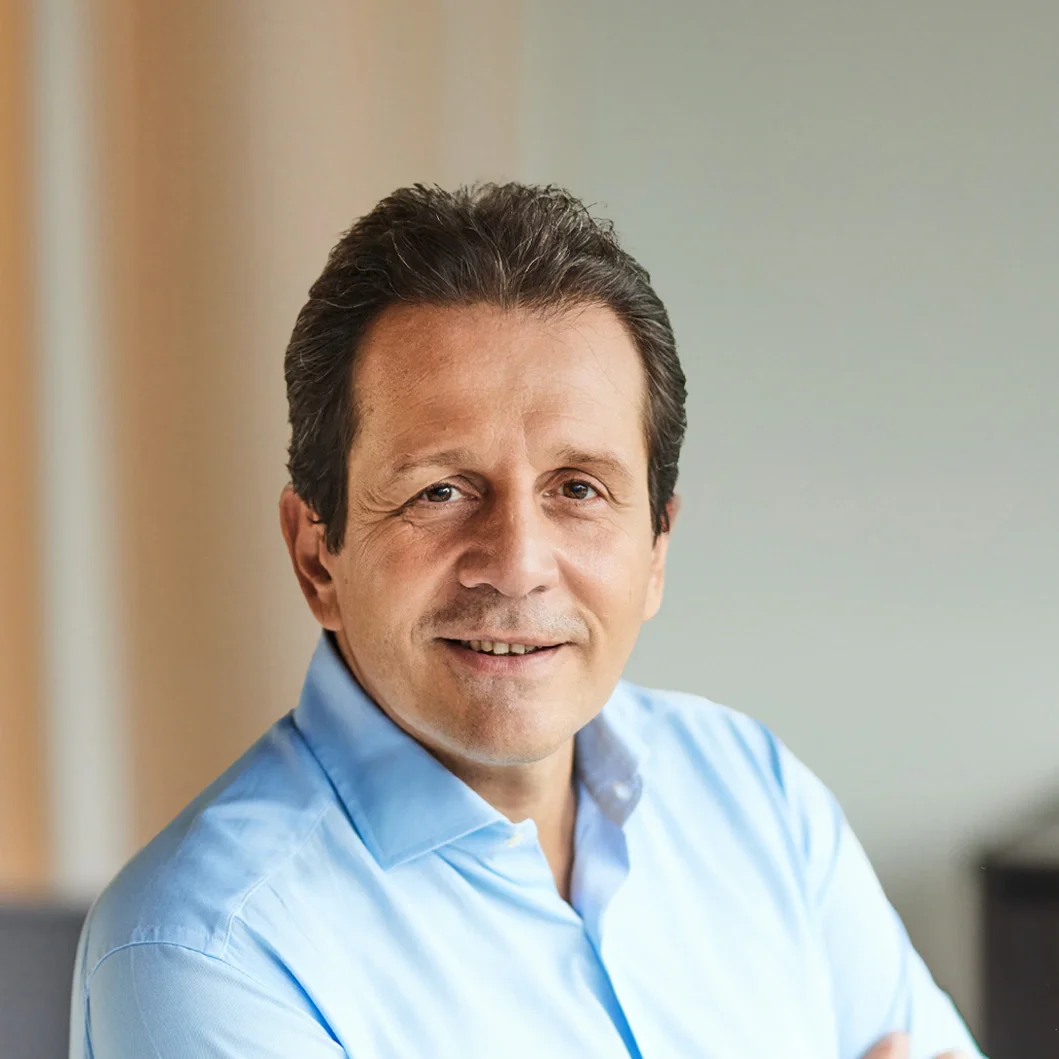 A smiling man with dark brown hair wearing a light blue shirt, sitting indoors with a blurred background.