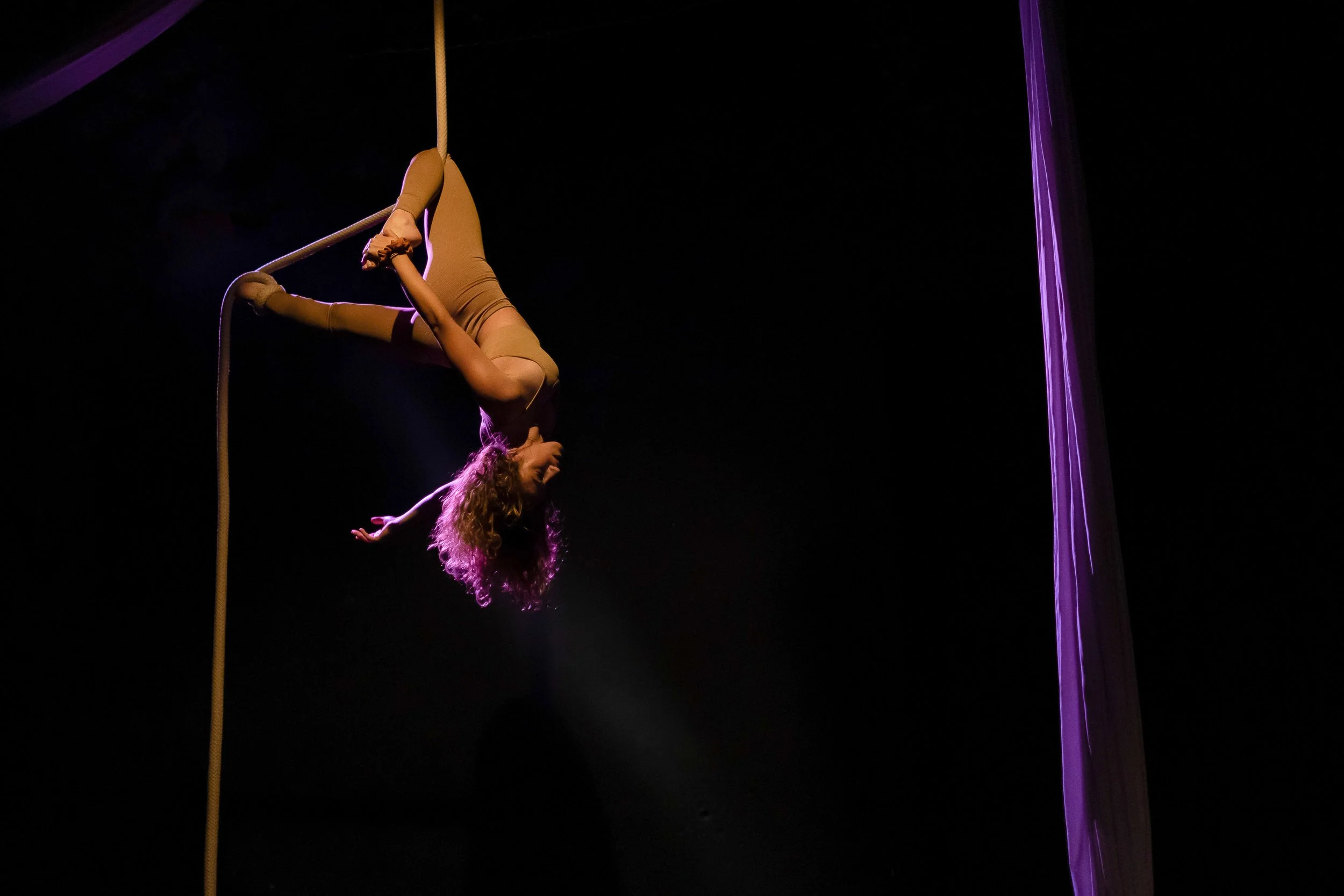 Una performer di lâ€™aerobica aerea che si esibisce sospesa in aria usando un attrezzo a forma di cerchio, con sfondo nero e illuminazione viola.