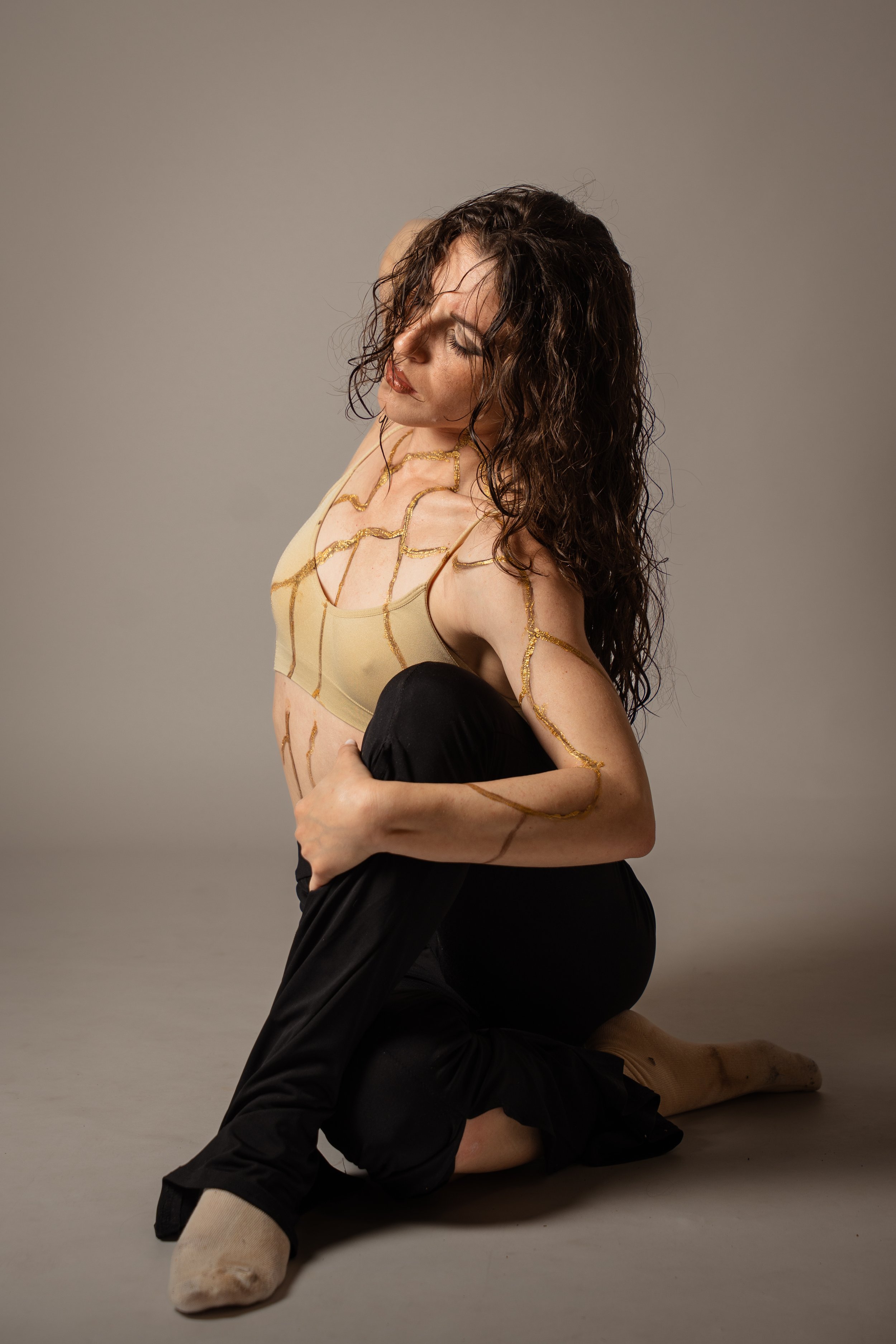 Donna con capelli ricci scuri e trucco neutro si inginocchia sul pavimento lucido, indossando un top beige con dettagli dorati e pantaloni neri, su sfondo grigio chiaro.