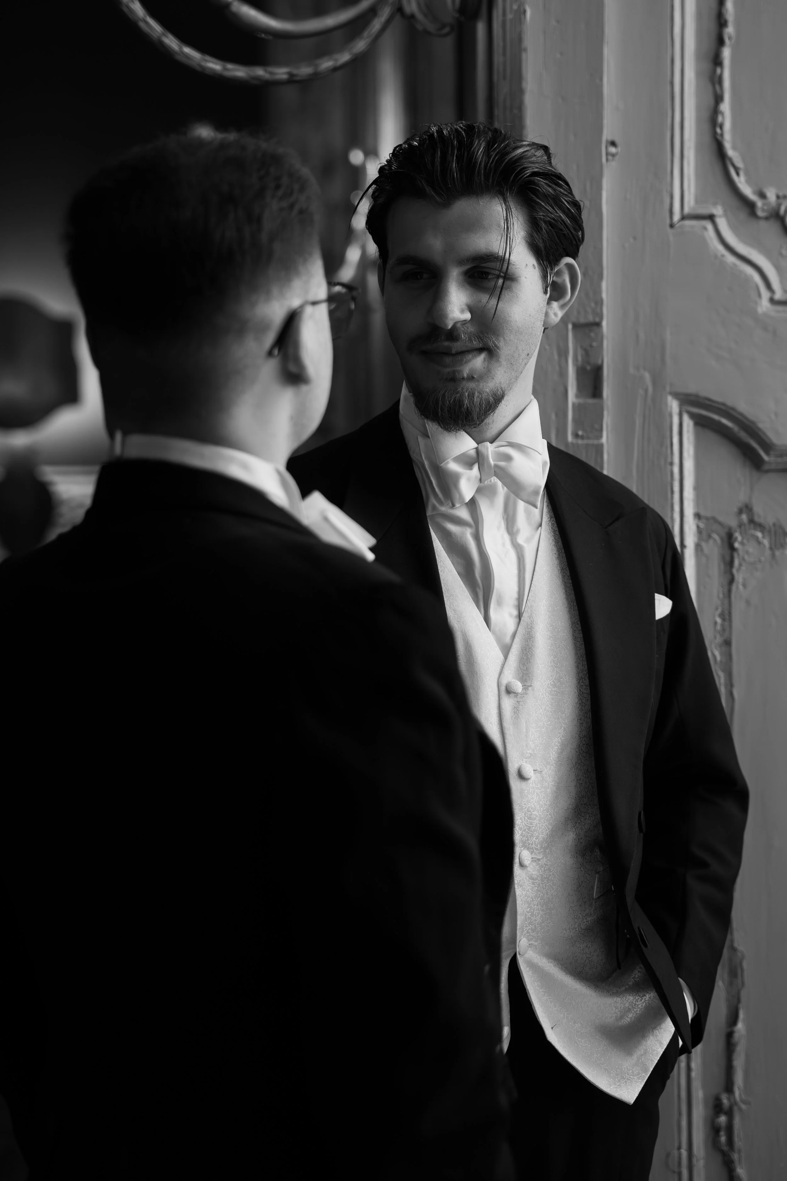 Due uomini eleganti in abiti da sera, uno con baffi e barba, si guardano mentre si conversano in un ambiente storico con pareti decorate.
